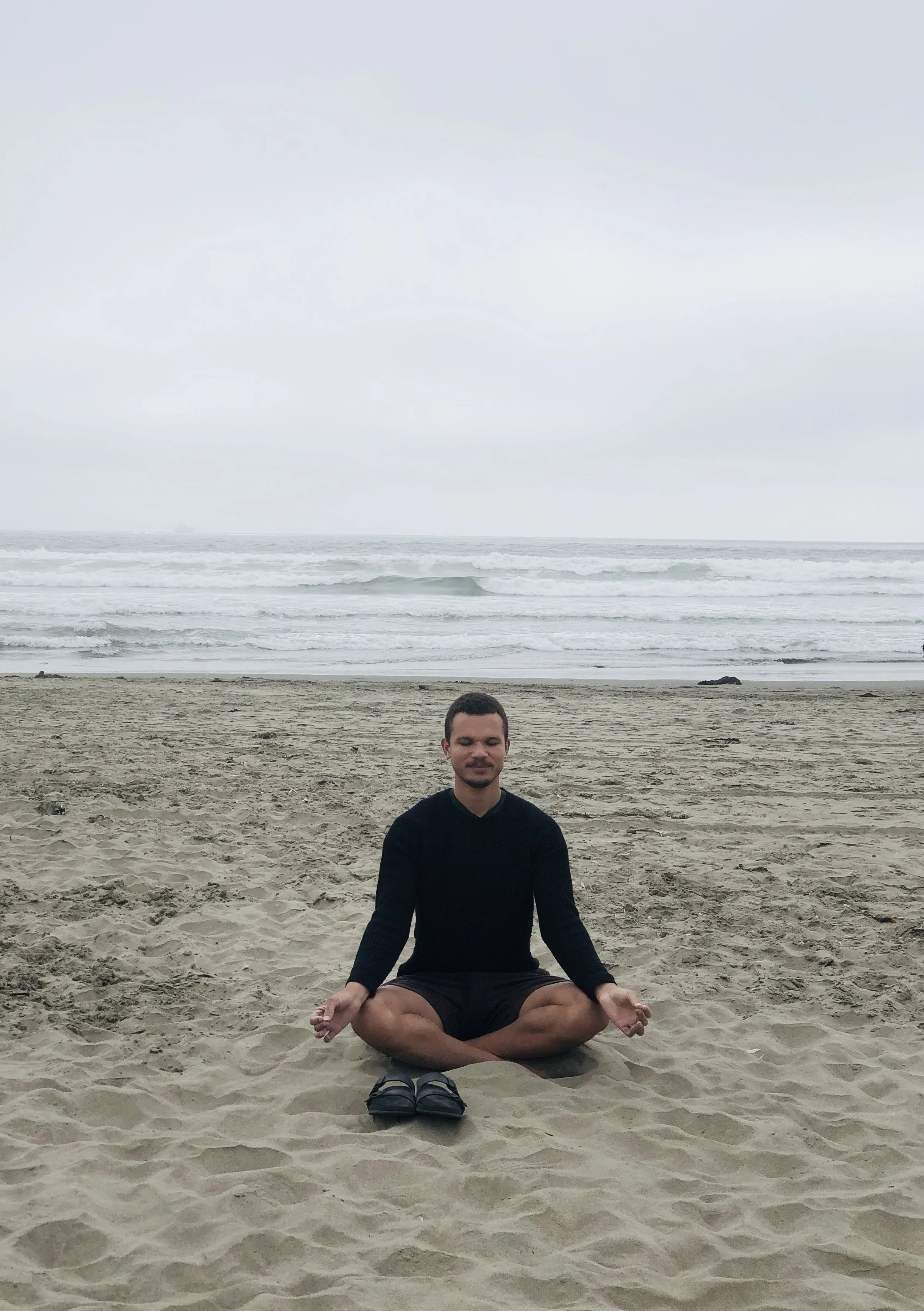 Ein Mann sitzt in einer Meditationspose im Schneidersitz auf einem Strand, mit dem Meer im Hintergrund, an einem bewölkten Tag.