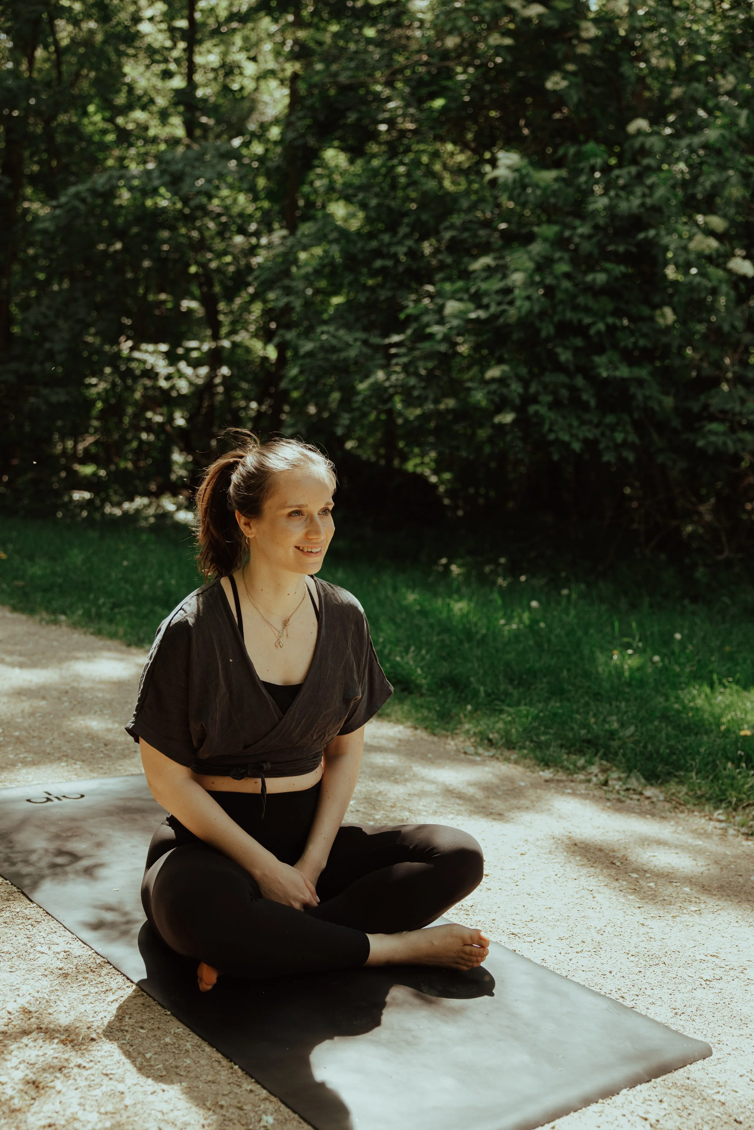 Junge Frau sitzt im Schneidersitz auf einer Yogamatte im Grünen, lächelt und praktiziert Yoga im Freien an einem sonnigen Tag.