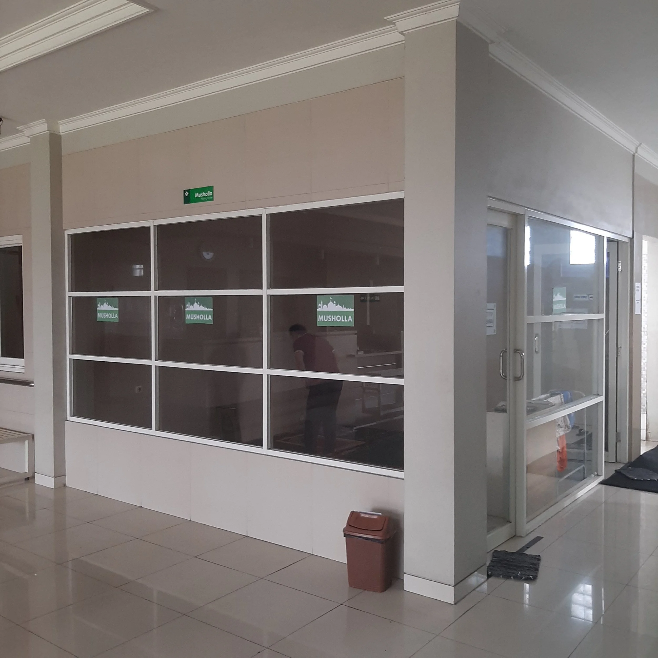Interior of a building with glass enclosures labeled "Musholla" for prayer rooms, with a trash bin and a person visible inside the room.
