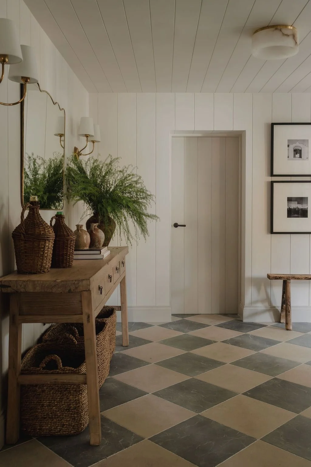 Interior hallway with a wooden console table decorated with vases and a large plant, black and white framed photographs on the wall, and a checkered tile floor.