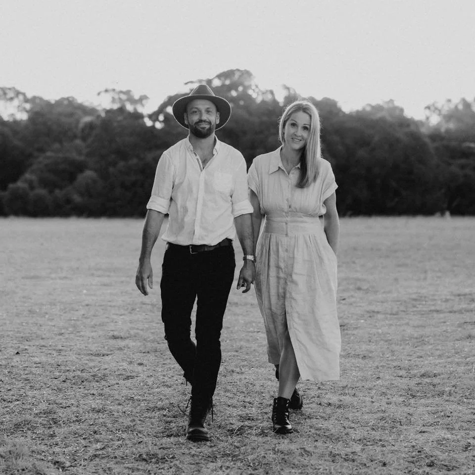 A man and woman walking hand in hand outdoors in a field, with trees in the background, in black and white.