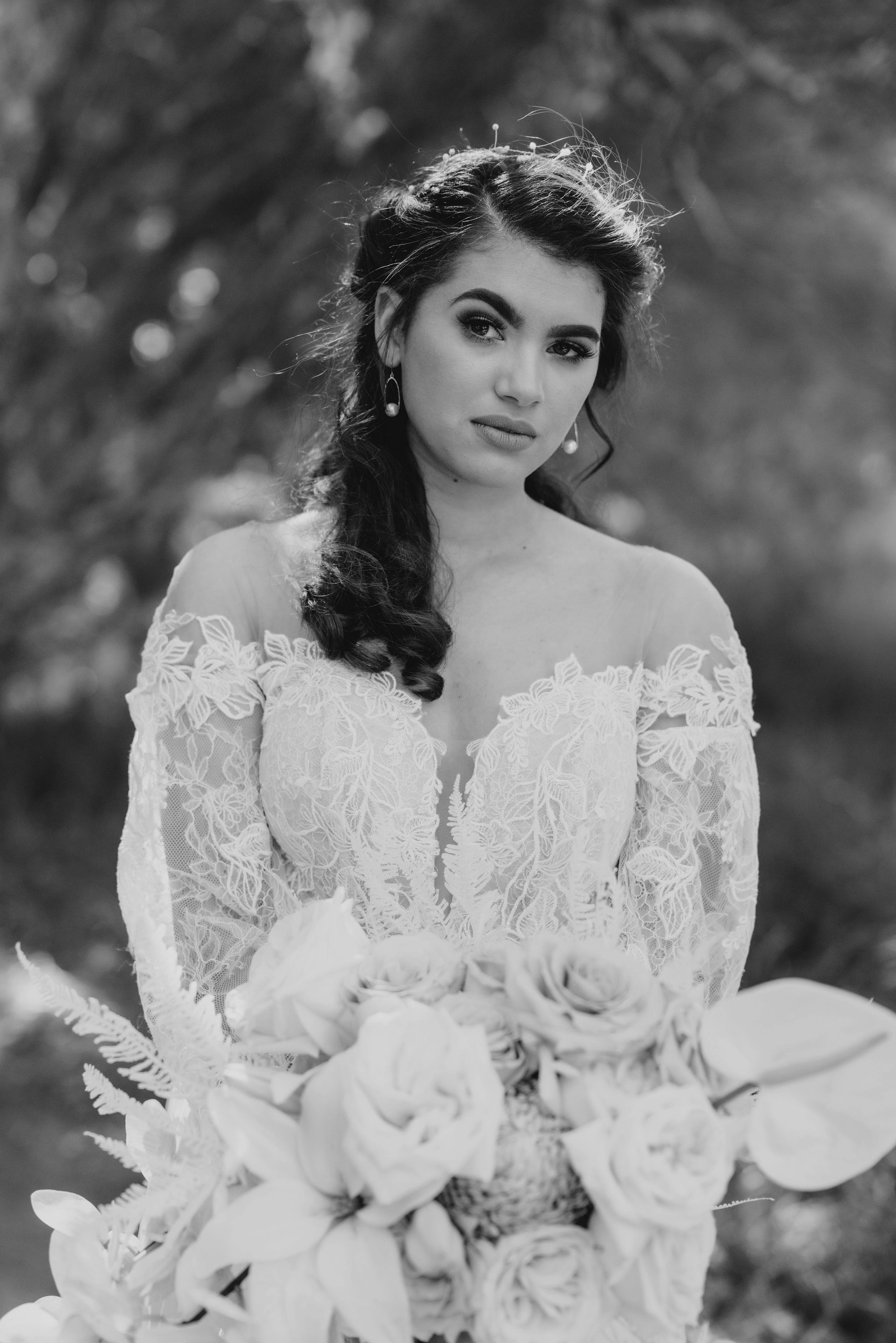 A bride in an off-the-shoulder lace wedding dress with long dark hair and earrings, holding a large bouquet of roses and other flowers, outdoors with blurred trees in the background.