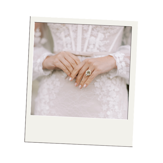 Close-up of a woman's hands with a large ring on her left middle finger, resting on her lap in a white lace dress.
