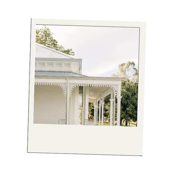 A white house with a detailed porch and a metal roof, surrounded by trees under a cloudy sky.