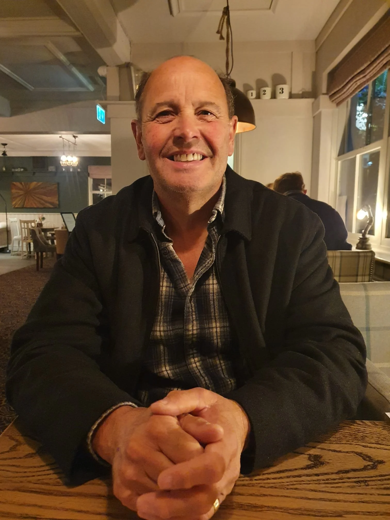 A middle-aged man smiling, sitting at a wooden table with his hands clasped in front of him. He has a receding hairline, wears a dark jacket over a checkered shirt, and is in a cozy interior with warm lighting, curtains, and framed artwork.
