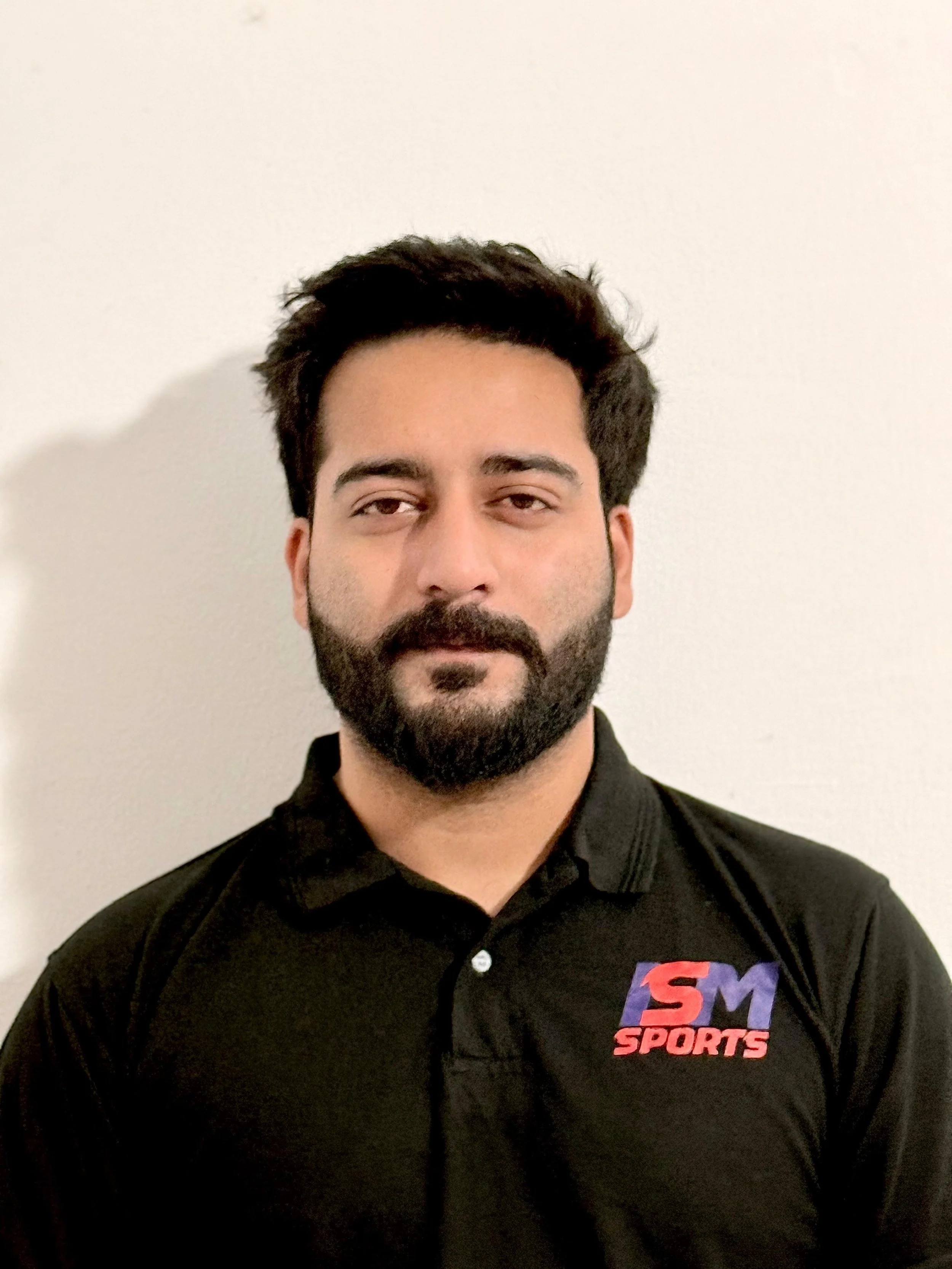 Man with a beard wearing a black polo shirt with "ISM Sports" logo, standing indoors, with tattooed right arm and neutral expression.