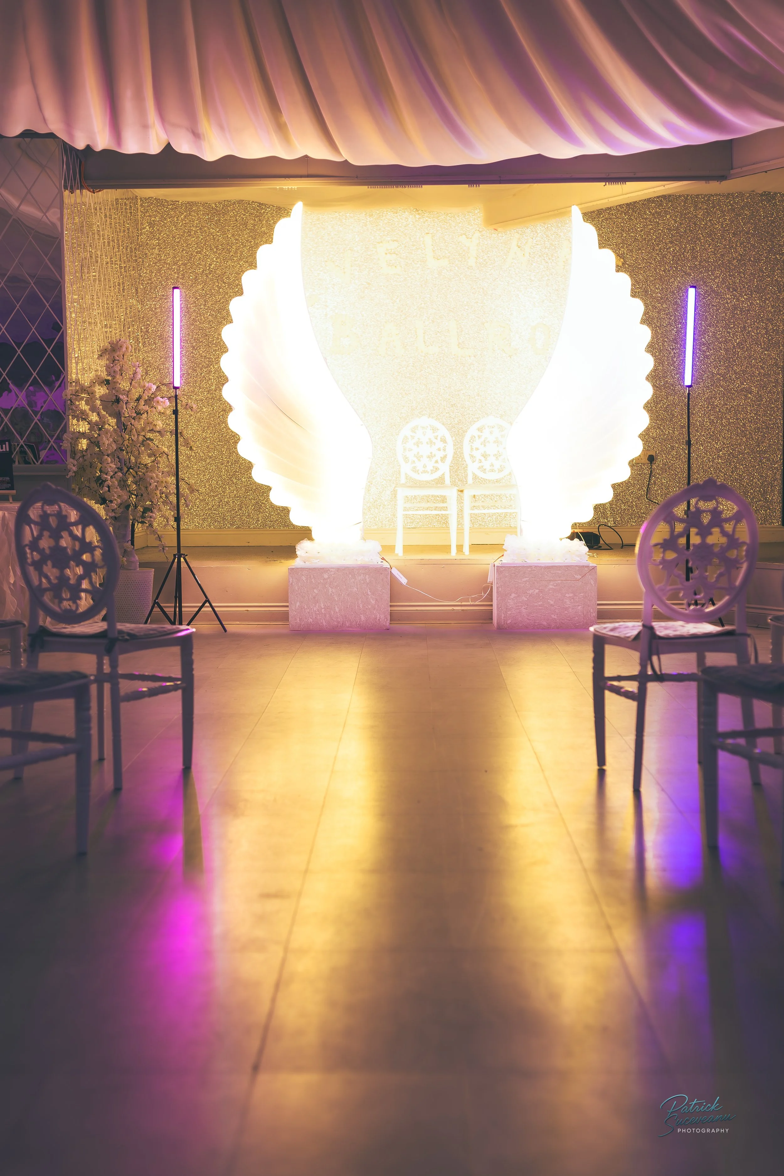Decorated event stage with illuminated angel wings and a glittery backdrop.