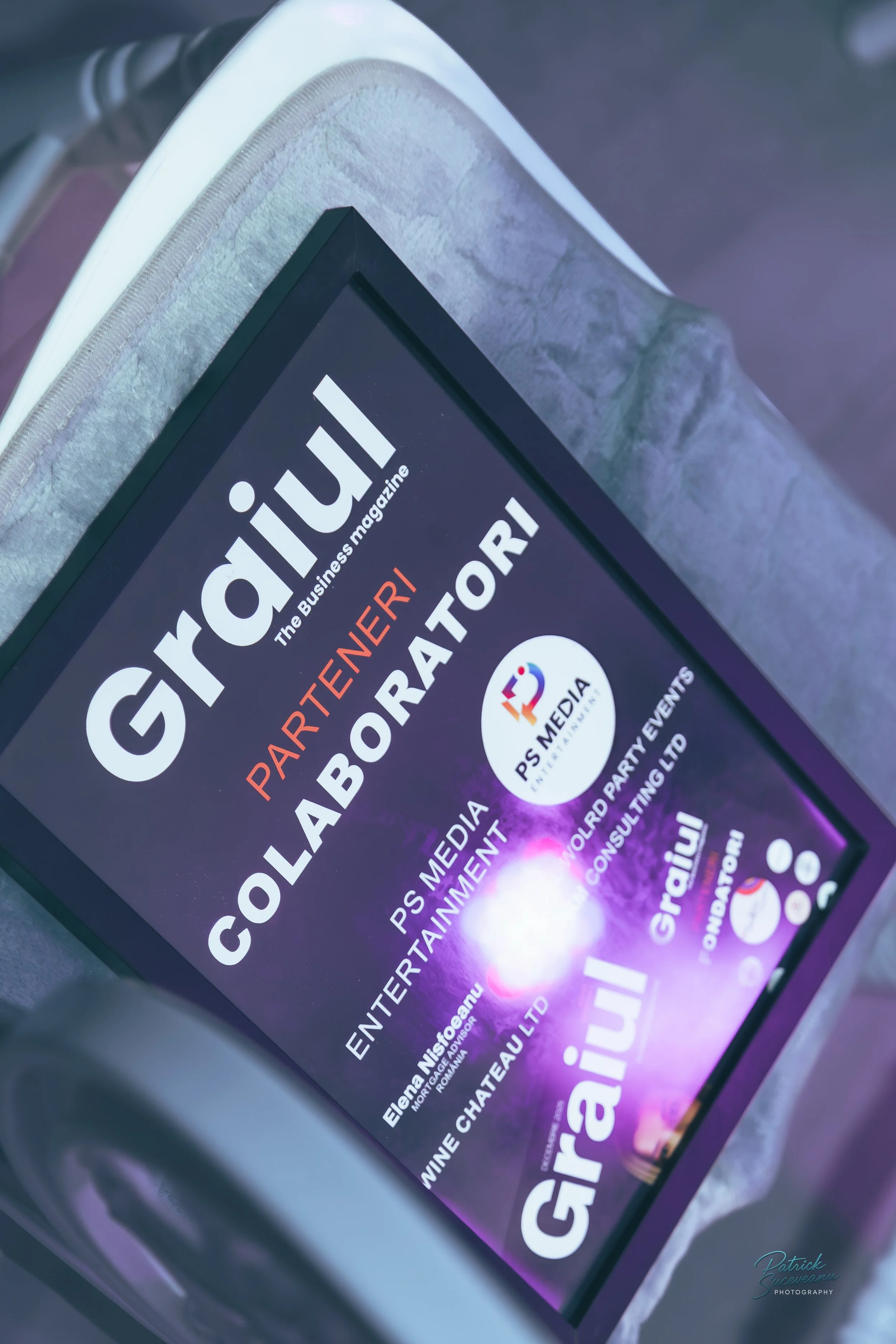 Digital screen displaying an event or partnership confirmation for Graiul, a business magazine, and PS Media Entertainment, with logos and event details, resting on a table with a textured silver cover.