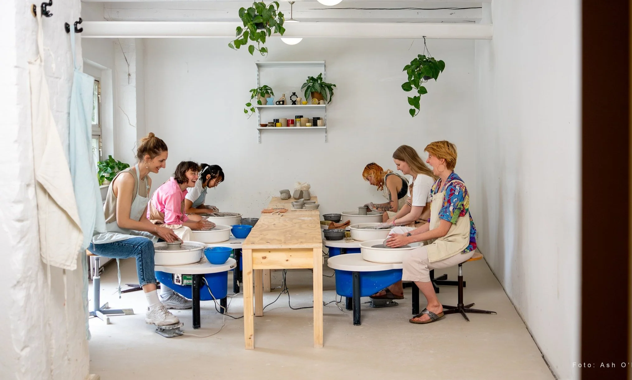 A group of women participating in a pottery class, working on pottery wheels in a bright, minimalist studio with plants and shelves on the white walls.