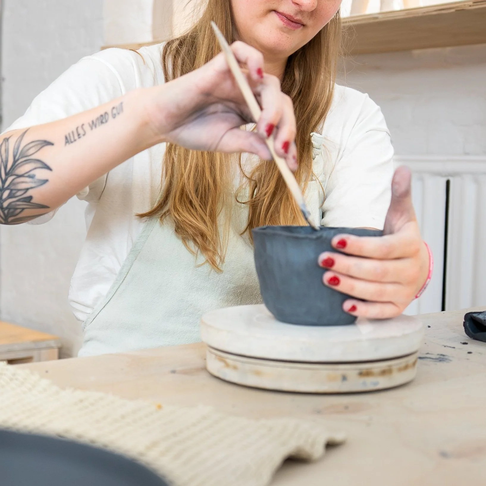 Person with red hair, tattooed arm, and red painted nails decorating a black ceramic bowl with a paintbrush in a light-colored room.