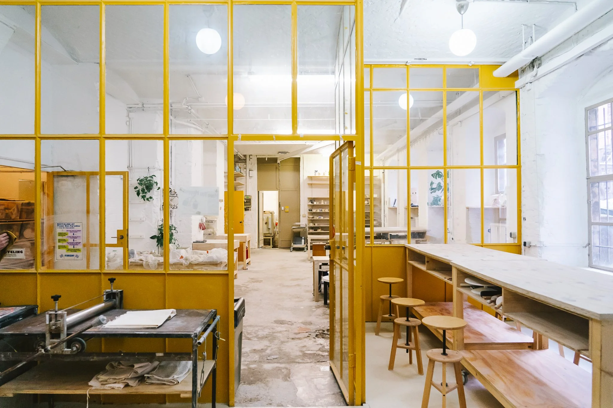 Interior of a modern office or coworking space with white walls, large windows, and yellow metal-framed glass partitions. There are wooden work tables with stools, some greenery, and minimal decor. Bright lighting highlights the open and airy layout.