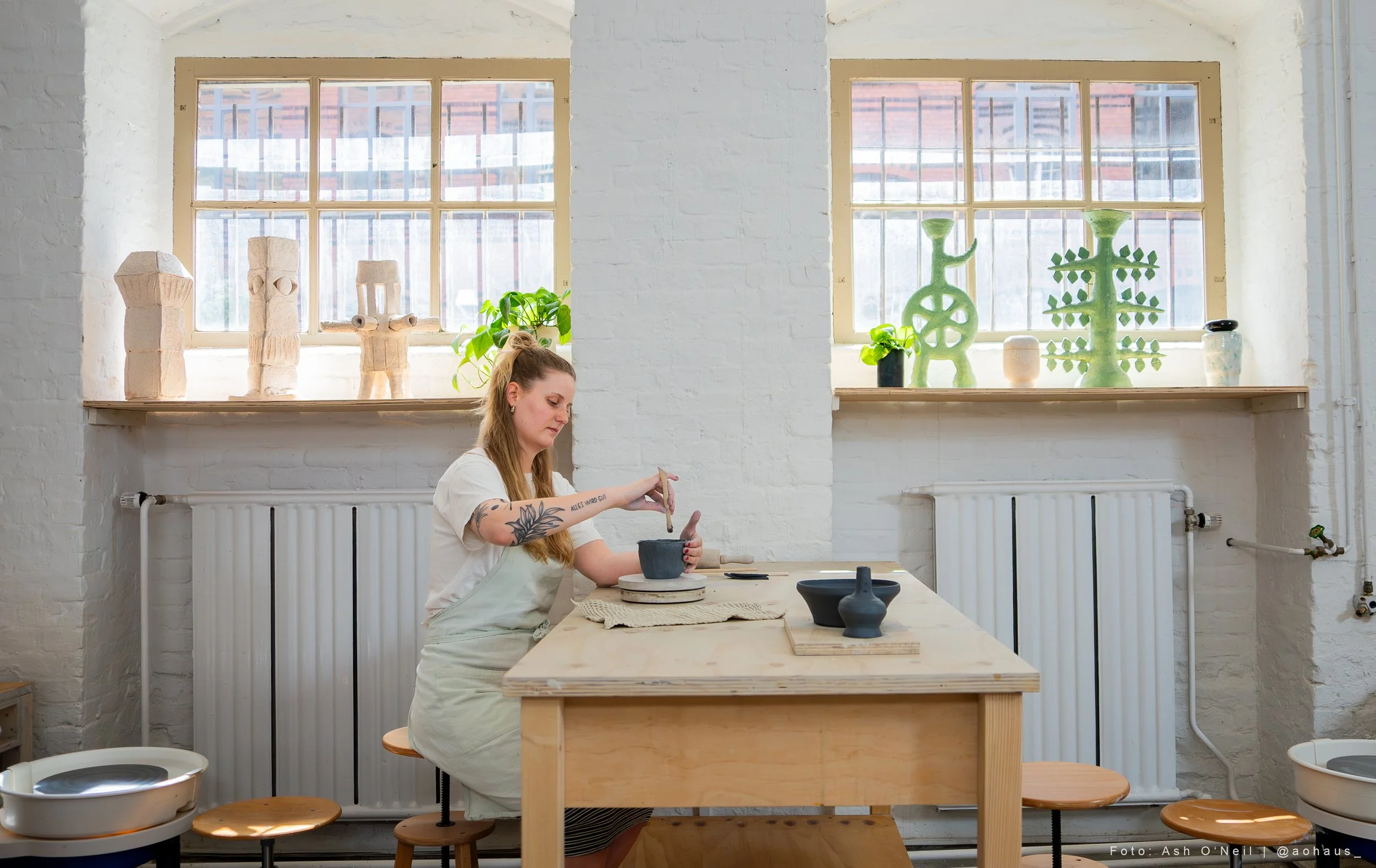 A woman working on a pottery project in a bright studio, sitting at a wooden table with pottery tools and black pottery pieces, with shelves of pottery and decorative sculptures behind her.
