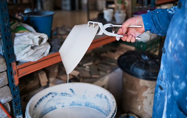 a person holds a ceramic bowl with glazing tongues. Glaze drips from the bowl into a large bucket of glaze