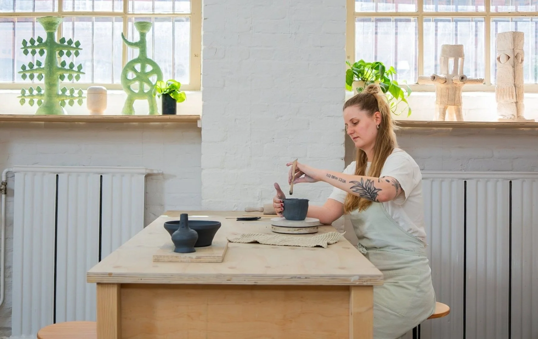 A woman with tattoos on her arm sitting at a wooden table in a bright, modern studio, sculpting a black ceramic bowl with a tool during a pottery class. On the table, there are other black ceramic pieces and a beige cloth.