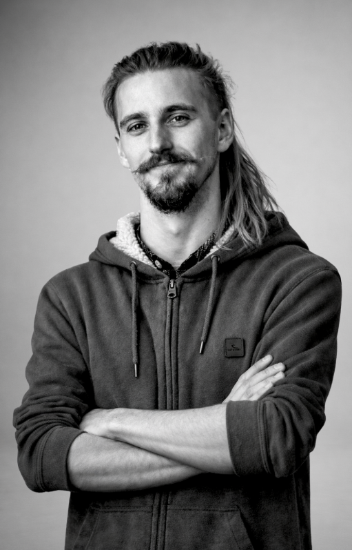 Black and white portrait of a man with long hair, beard, and mustache, wearing a hoodie with arms crossed, smiling slightly against a plain background.