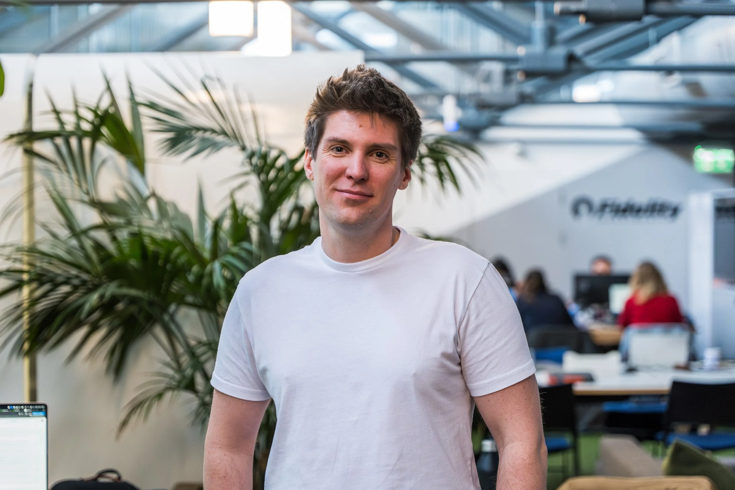 Man in white T-shirt standing indoors with plants and an office setting in the background.