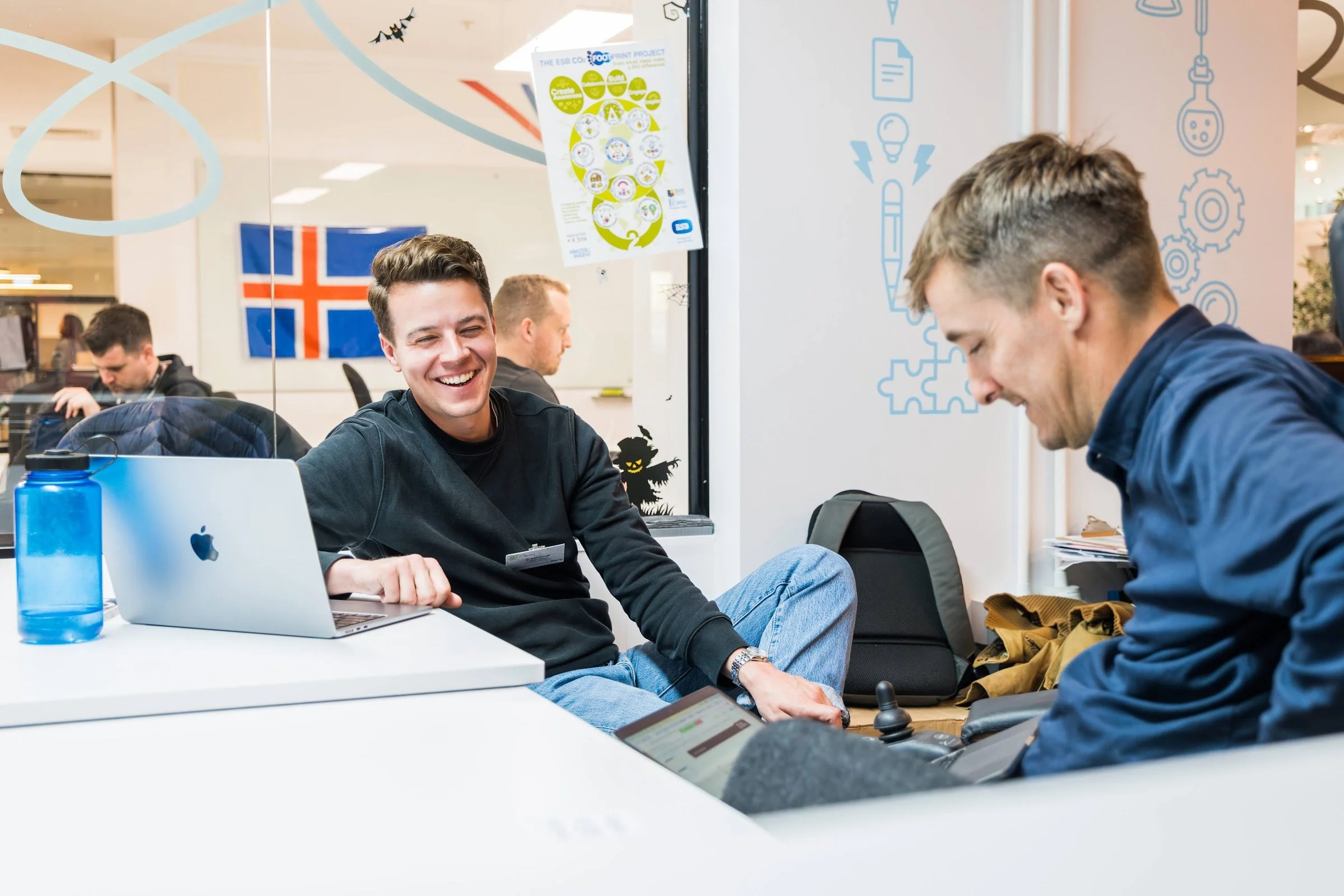 Two people sitting at a desk with laptops, smiling and talking, in an office with an Icelandic flag in the background. A blue water bottle is on the table.