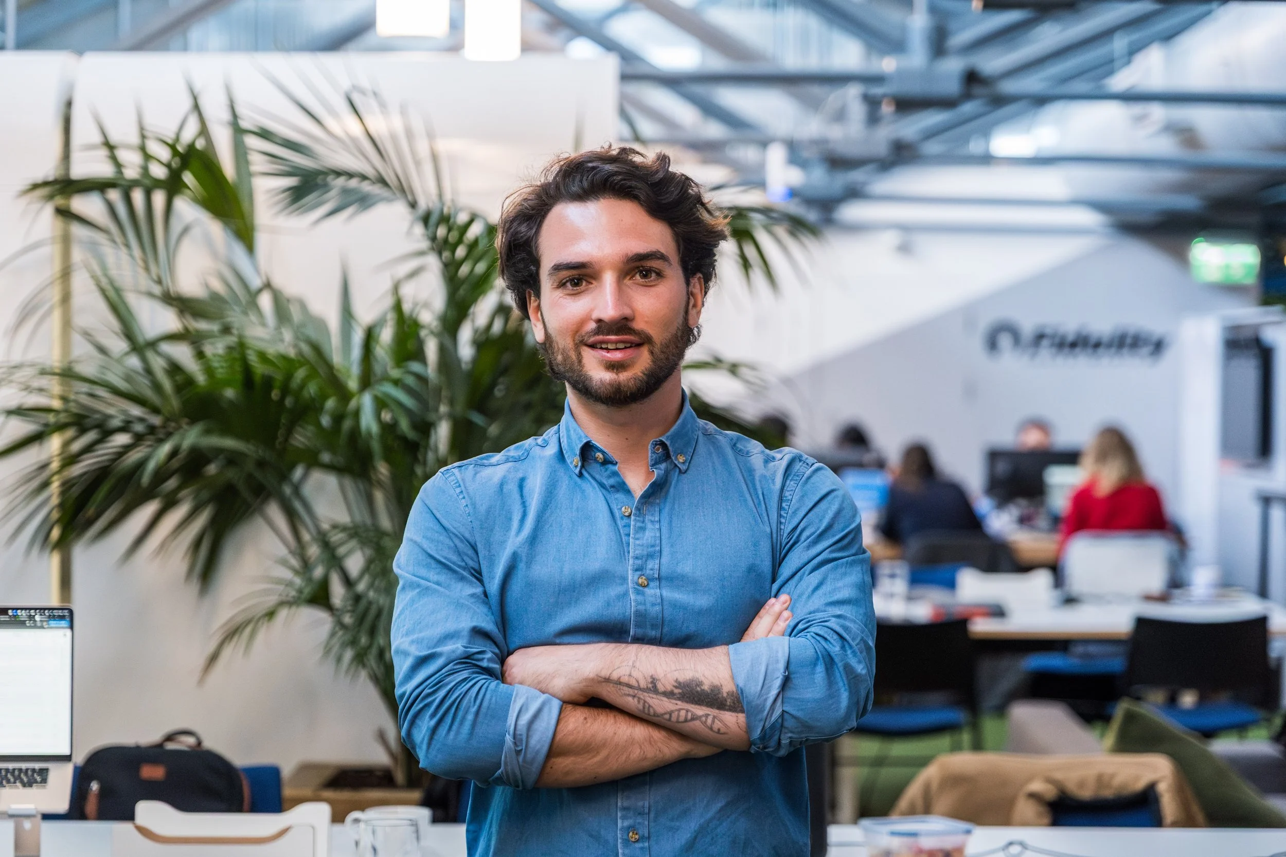 Person in a blue shirt standing with arms crossed in an office with plants and people working in the background.