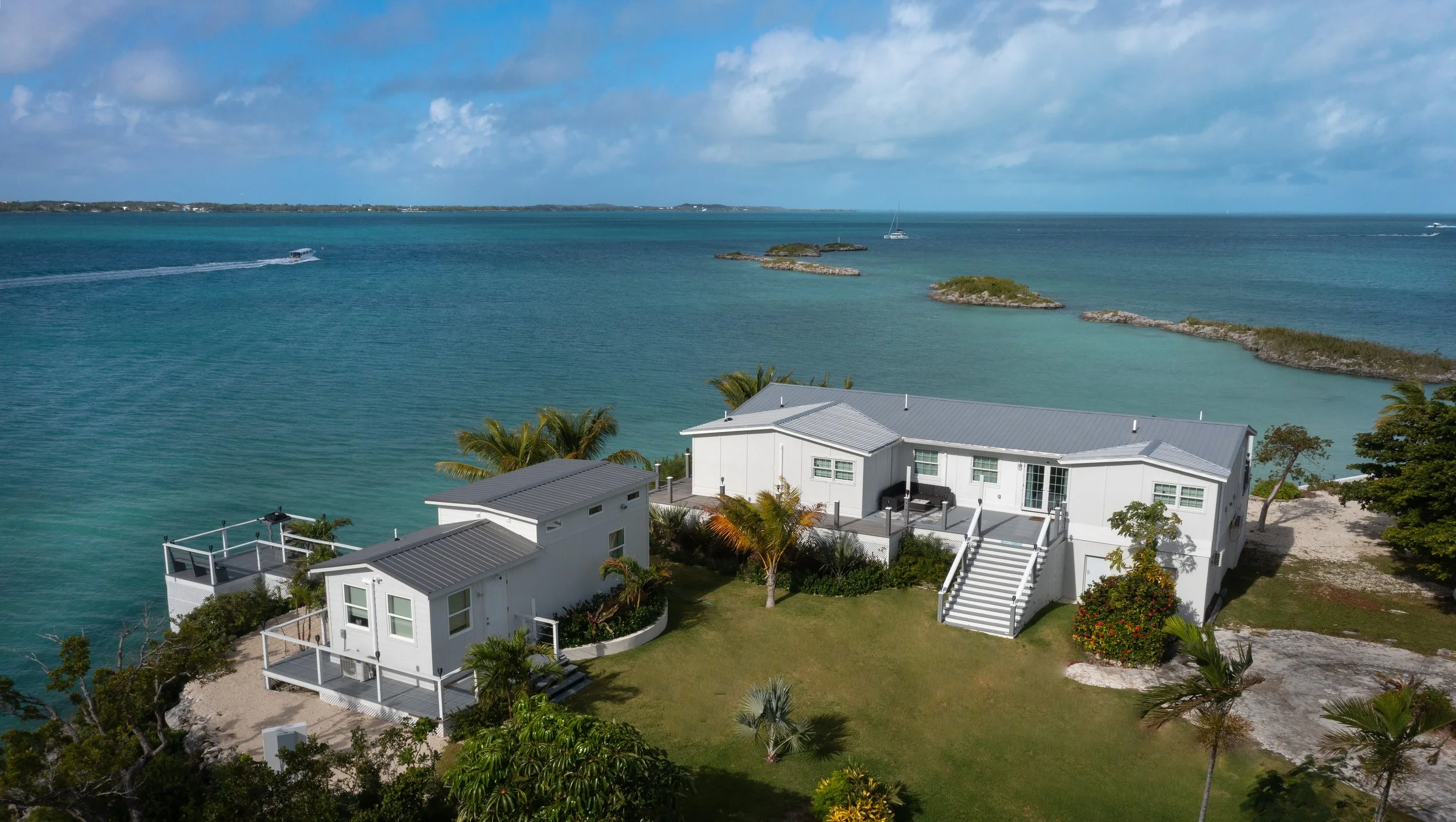 An aerial view of white beachfront houses with gray roofs on a lush green lawn, overlooking turquoise ocean water under a partly cloudy sky.