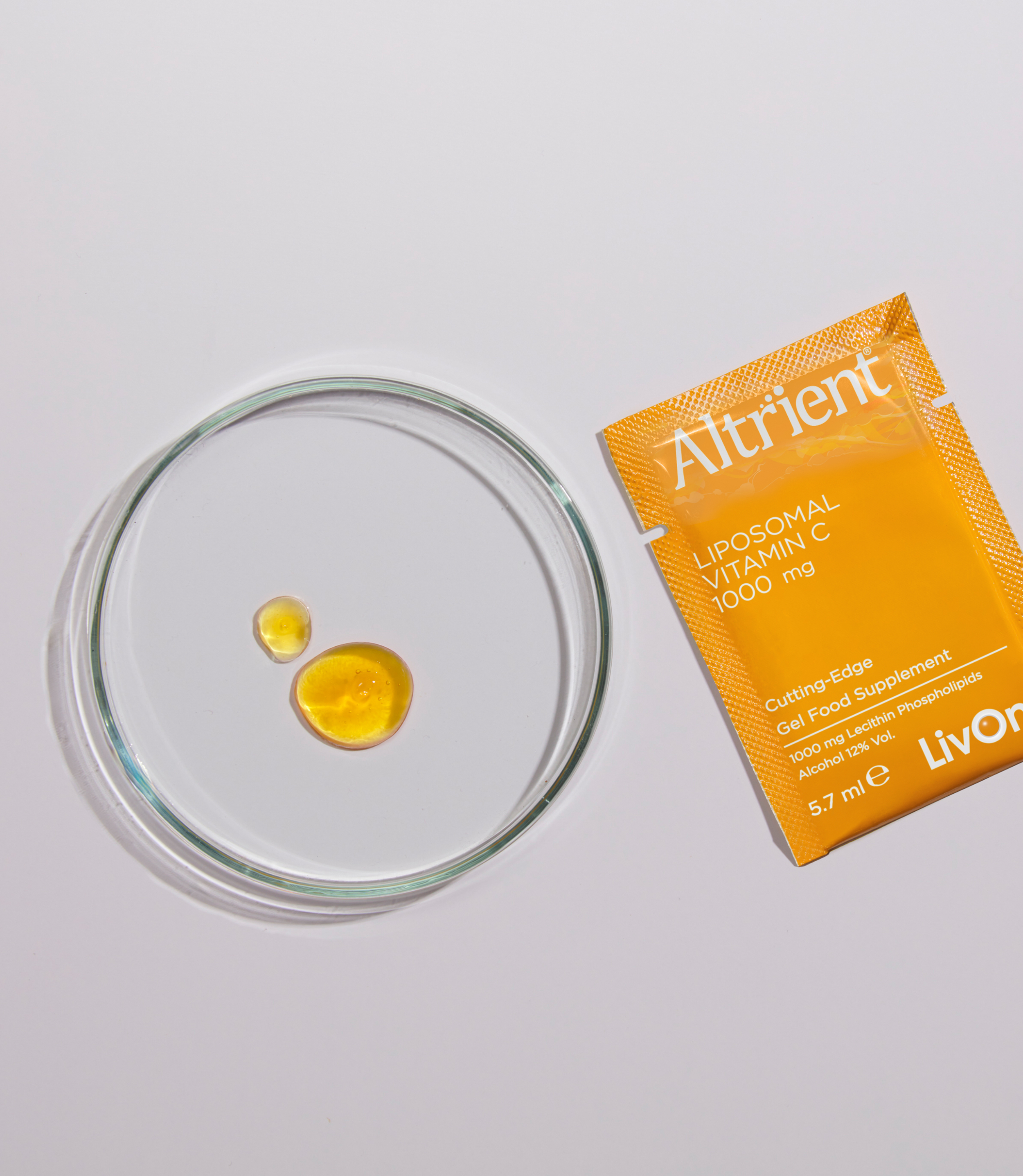 Two droplets of yellow liquid inside a clear glass petri dish and a packet of Altrient liposomal vitamin C on a white background.