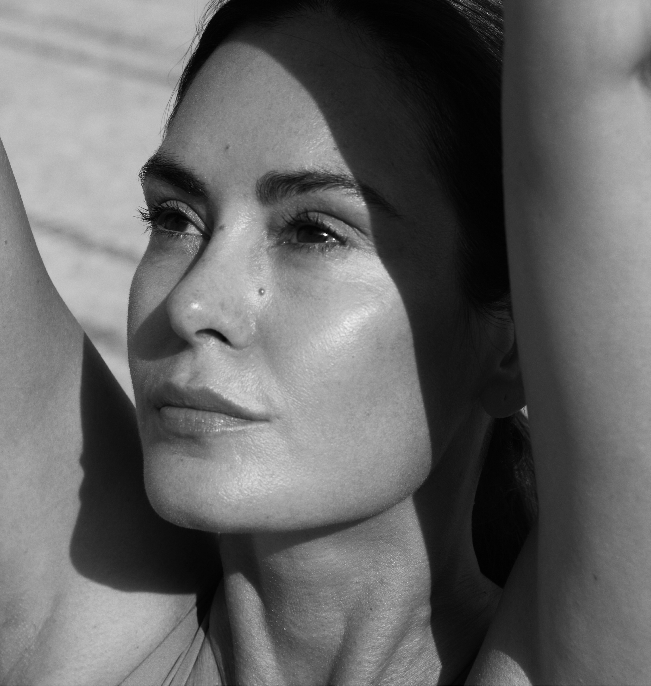 A black and white close-up photograph of a woman's face with her arms raised, casting a shadow across her face. She has a relaxed expression, visible pores, and a small nose piercing.