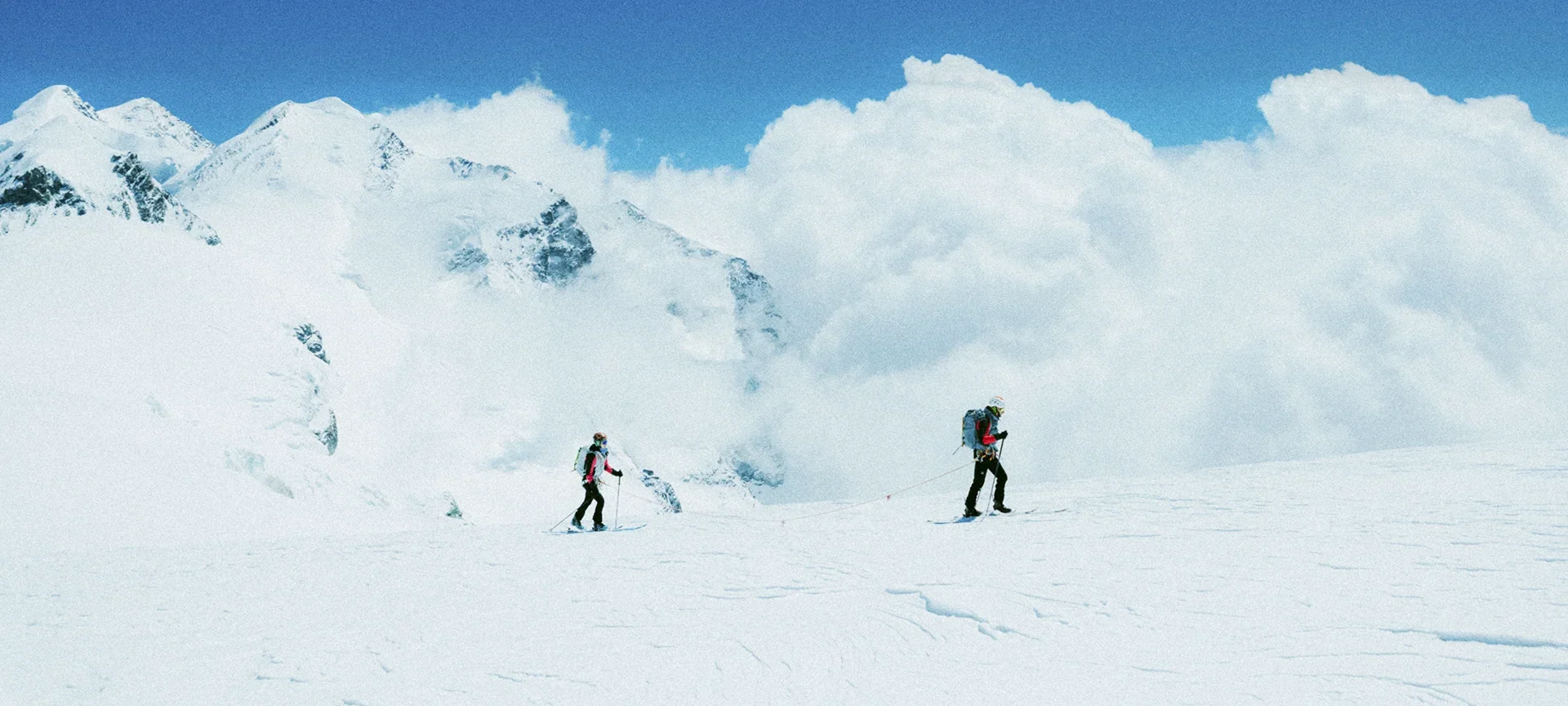 Display of a yellow ski jacket with red rope design, mounted in a glass case, against a background photo of skiers on a snowy slope, with timeline text indicating 2000 Old SkiSky and 2025 New SkiSky.