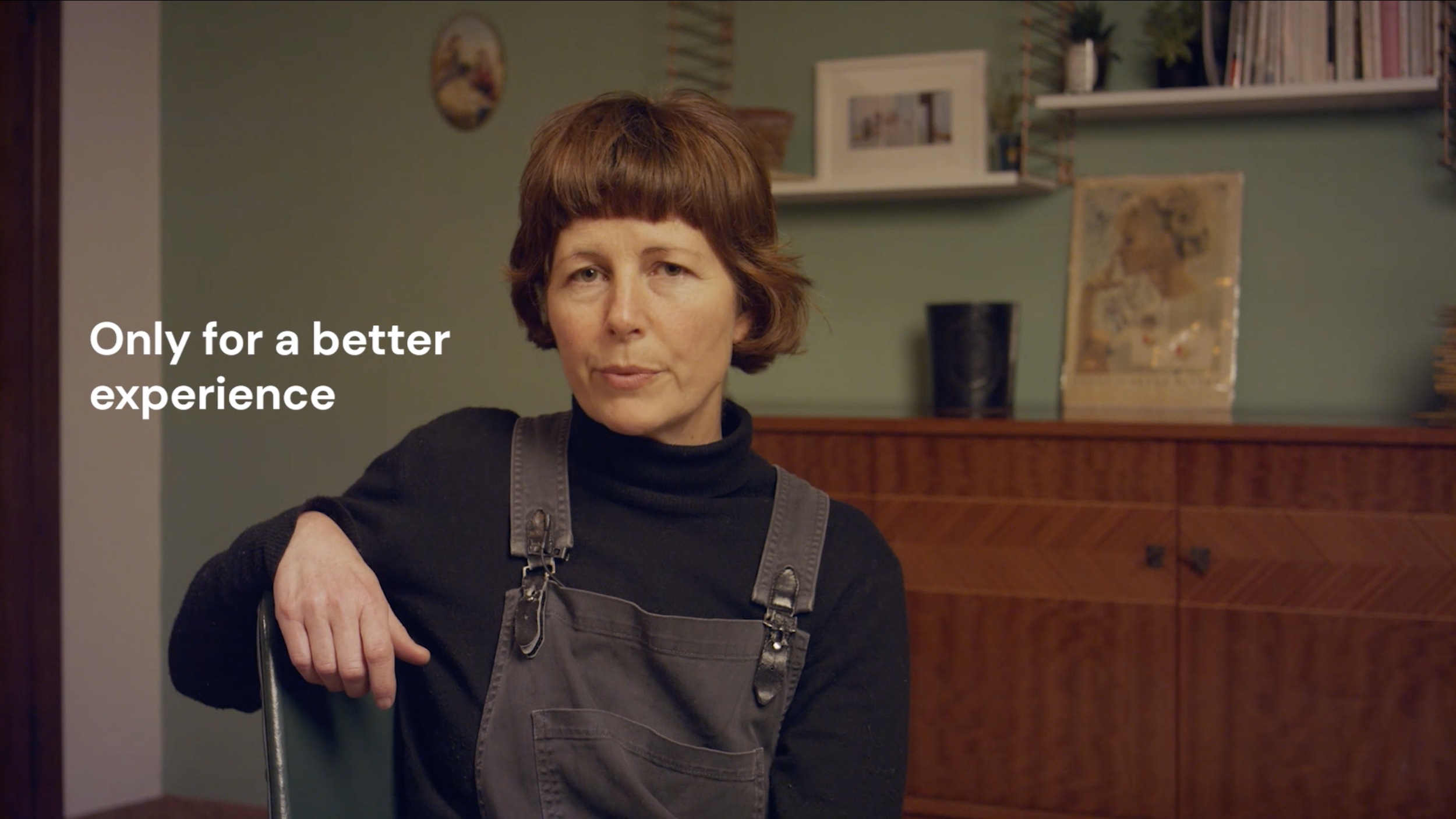 A woman with short red hair wearing a black turtleneck and gray overalls sits in a room with a wooden cabinet and shelves. The text on the image reads 'Only for a better experience.'