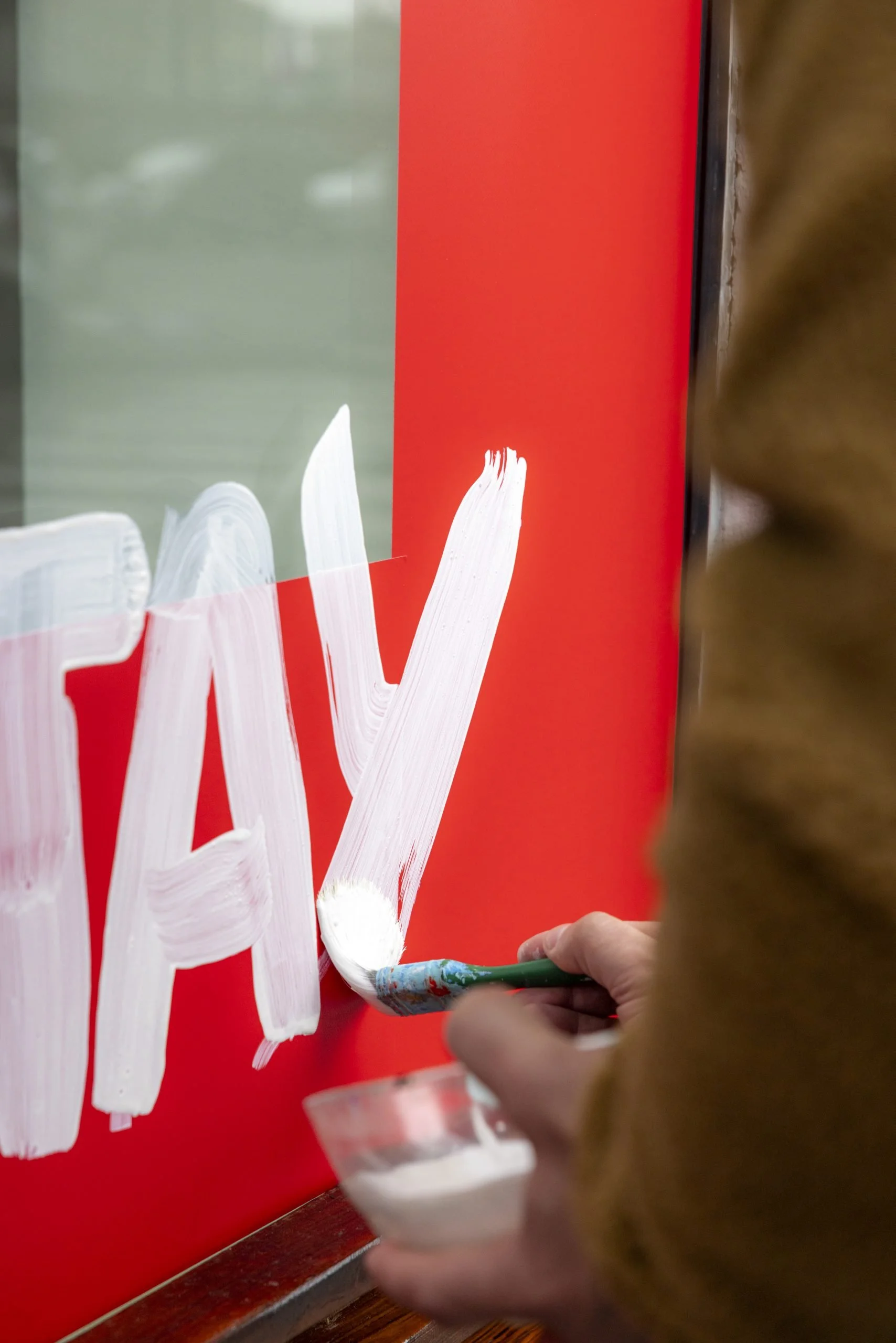 Close-up of a person painting white letters on a red surface with a brush, while holding a small container of white paint.