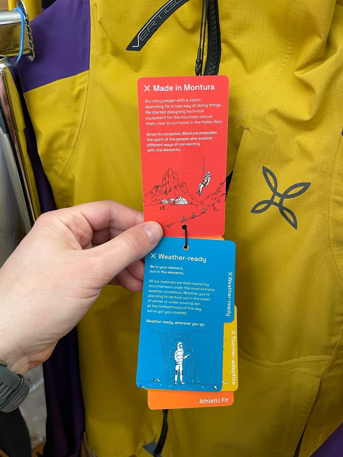 Two clothing tags attached to a yellow and purple outdoor jacket. The red tag discusses the brand's story and connection to Italian Alps. The blue tag highlights weather readiness and durability for extreme conditions.