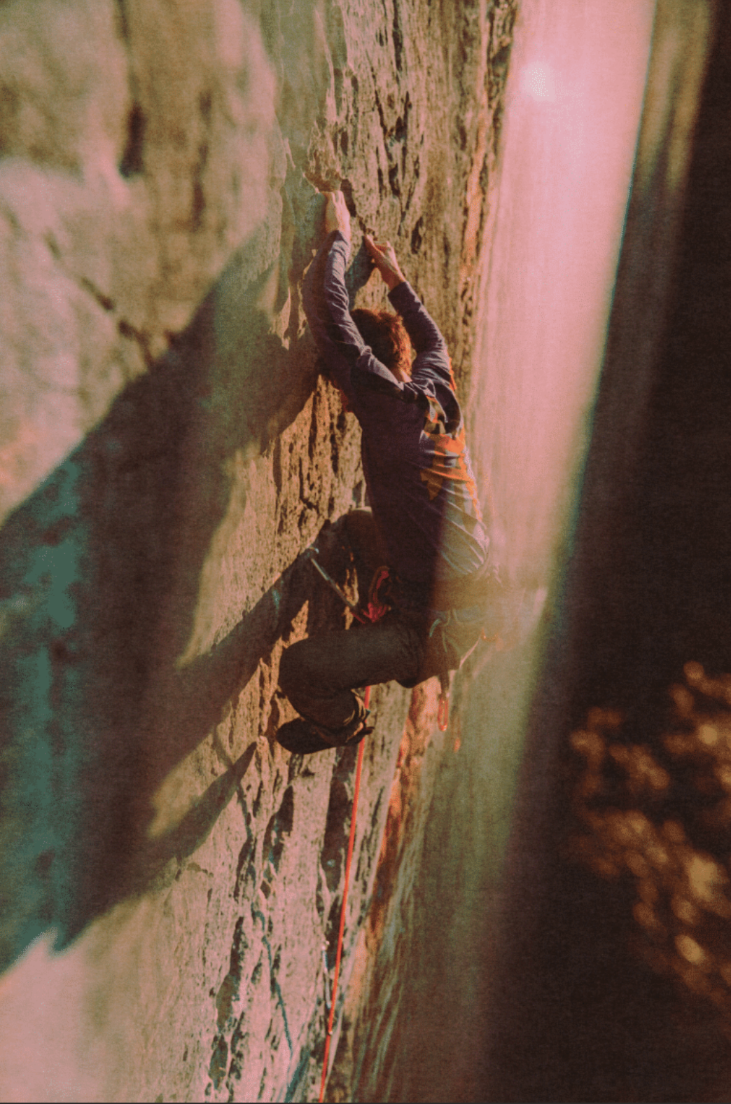 A person rock climbing on a steep outdoor cliff at sunset, using climbing gear and a red safety rope.