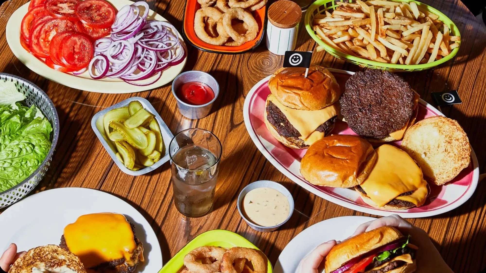 A variety of food on a wooden table including cheeseburgers, french fries, onion rings, sliced tomatoes, sliced onions, pickles, lettuce, ketchup, and dipping sauces.