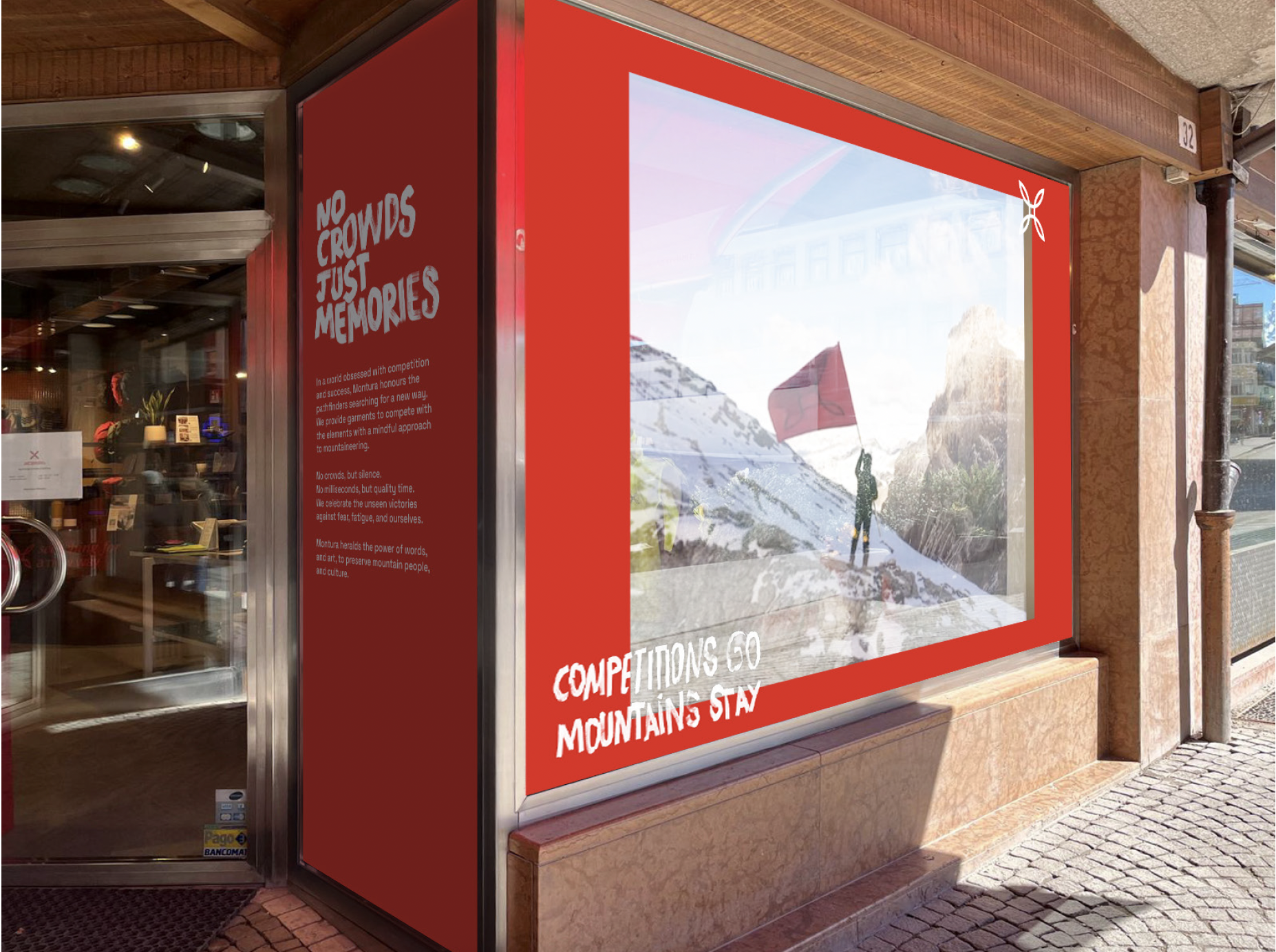A storefront window display with a red border showing a person holding a flag on a mountain with snow and rocky terrain. Text on the display says "COMPETITIONS GO MOUNTAINS STAY." Inside, through the glass door, there is a glimpse of the interior of a shop or cafe, with items on shelves and a potted plant visible.