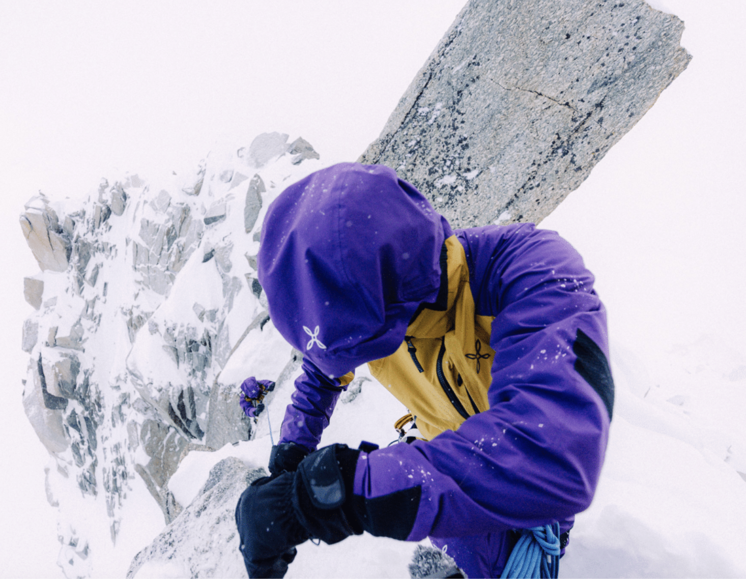 A mountaineer climbing a snowy, icy mountain with a large rock face behind him, wearing a purple and yellow mountaineering suit and gloves.