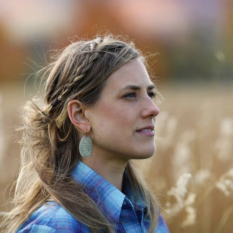 Hilary, a white woman with medium blonde hair and blue plaid button down shirt, looks out into a field of wheat.