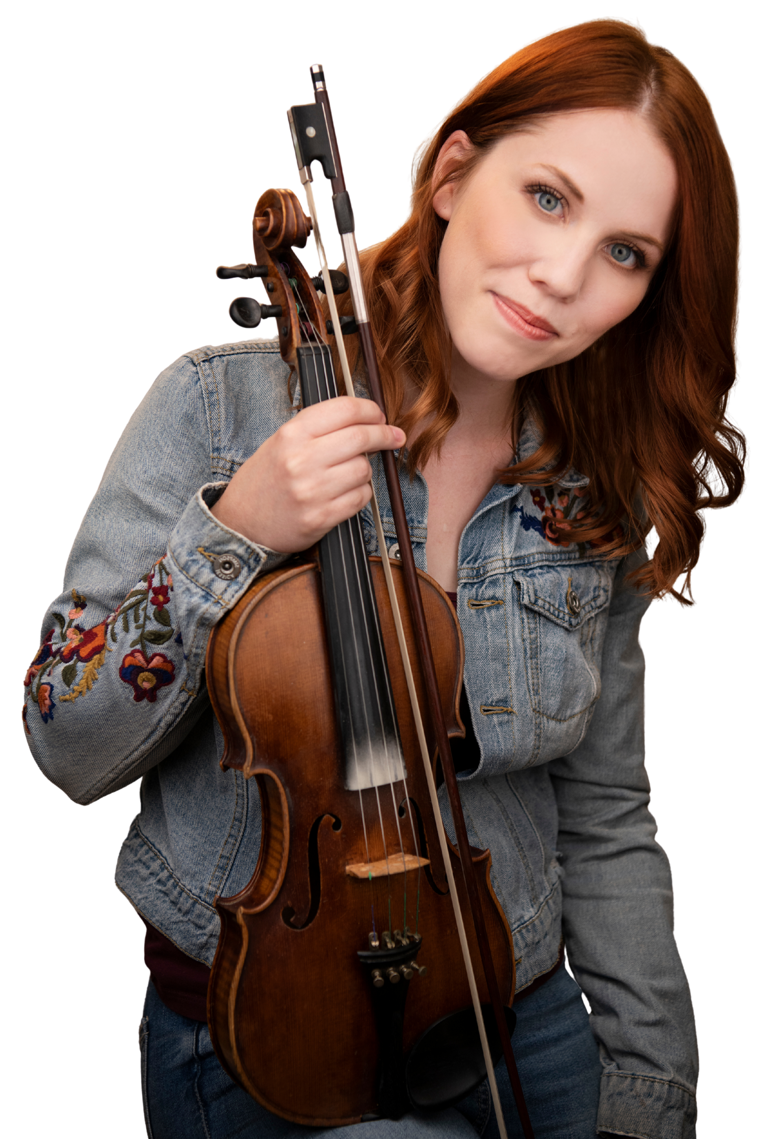 A white woman with long red hair smiles and wears a blue denim jacket. She tilts her head to one side and holds a fiddle and bow in one hand.