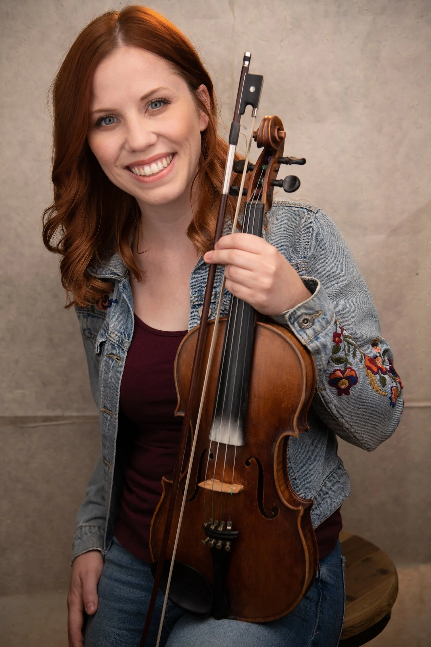Jo Brook- A white woman with medium auburn hair, blue eyes and a big smile, wears a jean jacket and holds her fiddle.