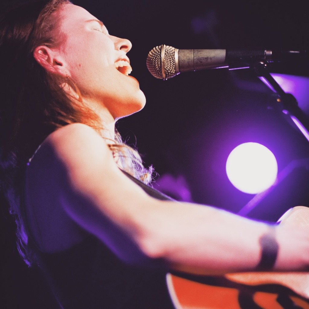 Brittany, a white woman with short brown hair sings into the mic while playing her guitar on stage.