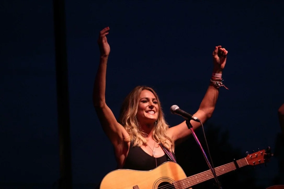Courtney, a white woman with long blonde hair and her guitar hanging in front of her, smiles with her arms up in the air.