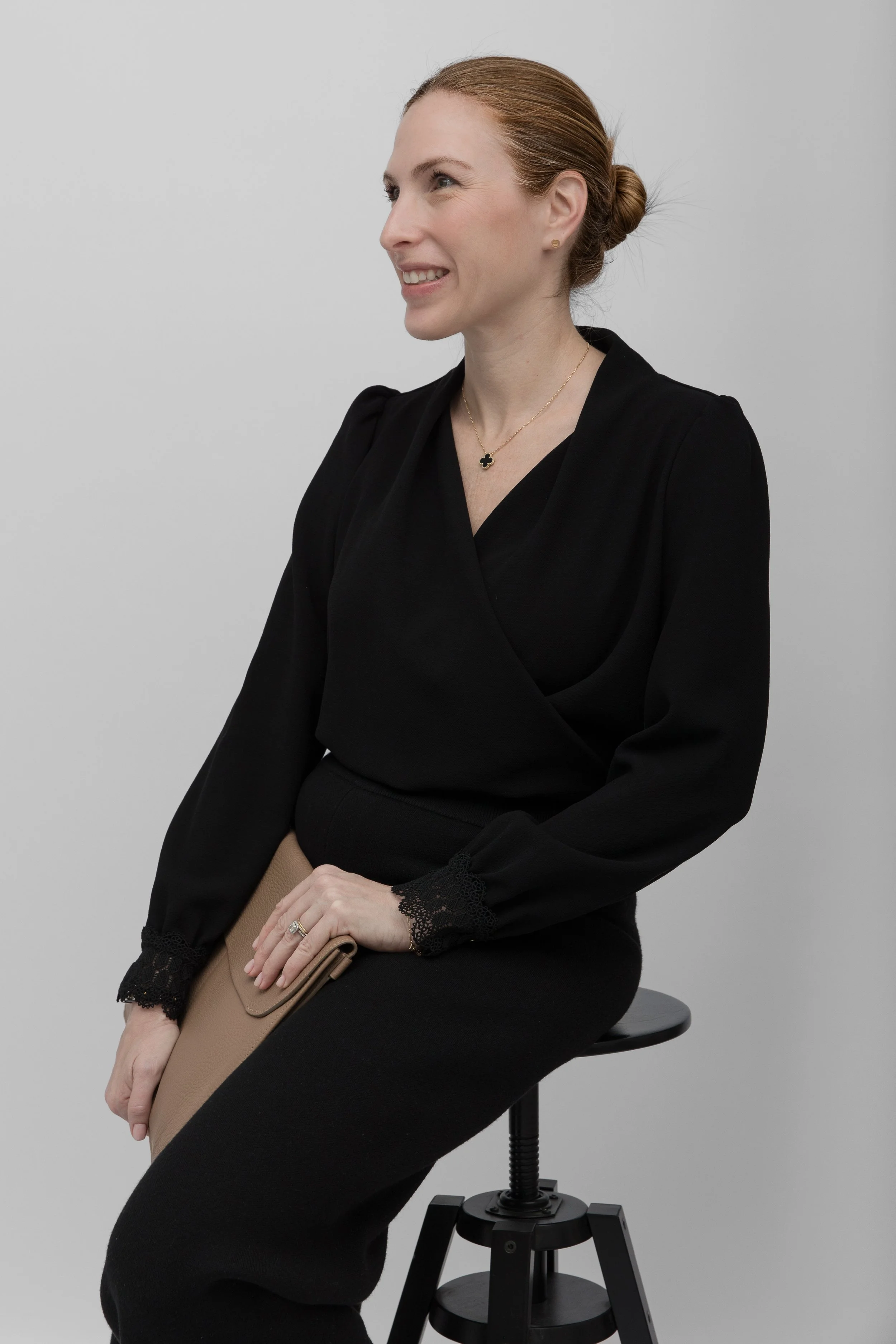 red hair tied in a bun, wearing a black long sleeve top with lace cuffs, and black pants, sitting on a black stool against a plain gray background. She is smiling, looking to her right, with a gold necklace with a black clover-shaped pendant and a ring on her left hand holding a folded tan clutch.