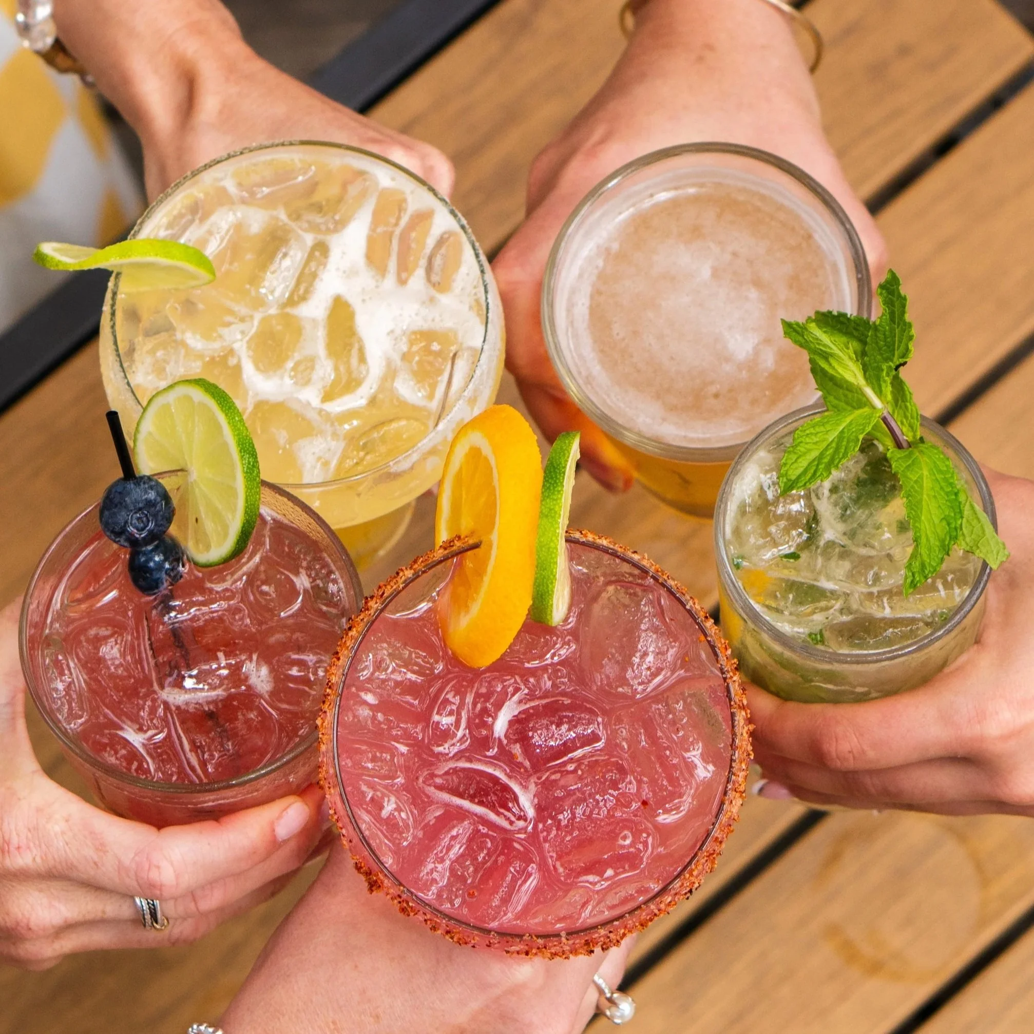 Top-down view of multiple colorful cocktails with garnishes, held in hands over a wooden table.
