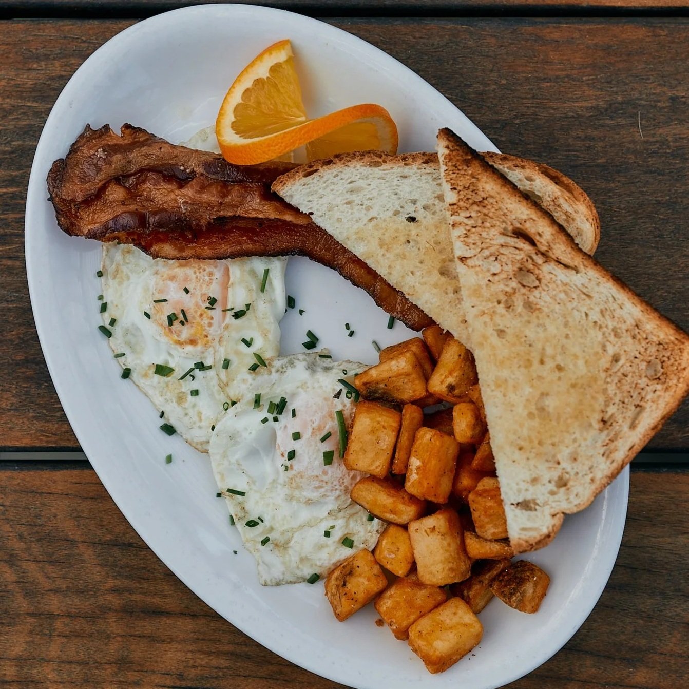 Breakfast plate with bacon, two fried eggs garnished with chopped chives, diced hash browns, toasted bread, and orange slices.