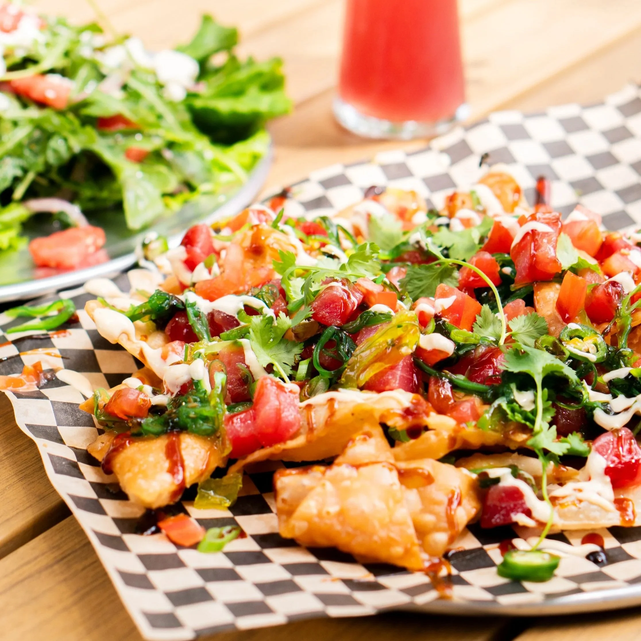 Close-up of tuna poke nachos on checkered paper with salad and a glass of pink drink in the background.