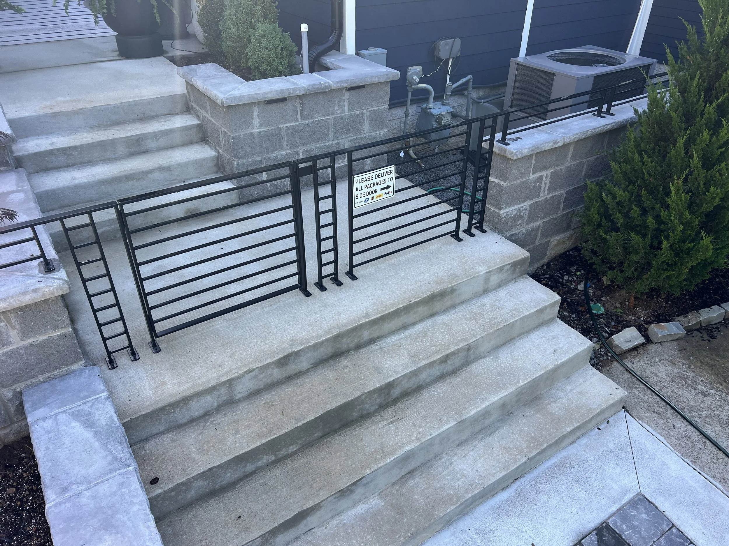 Concrete steps leading to a patio area with a metal gate that has a sign reading "Please deliver all packages to side door." The area is bordered by stone walls and landscaping, including bushes and a small garden bed.