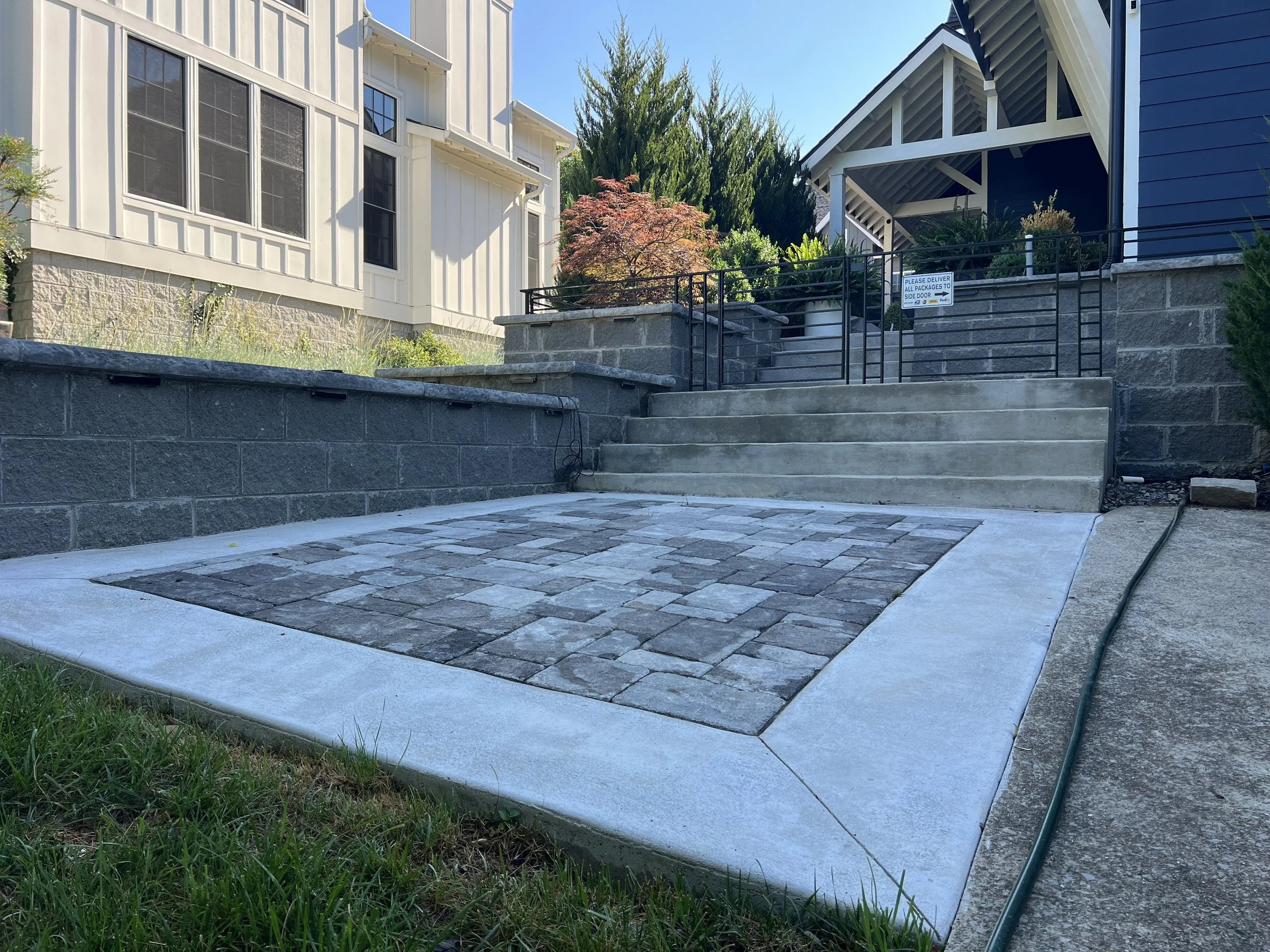 Backyard with stone patio, concrete stairs, black metal fence, and two houses with different siding styles.
