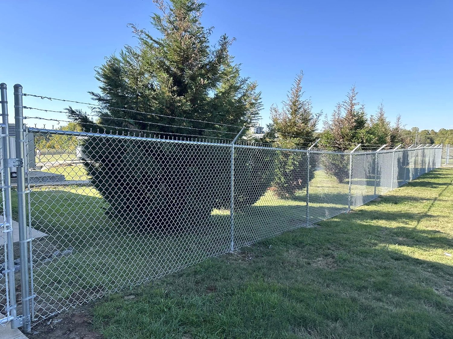 A chain-link fence built by Heritage Fence serving Paducah, Murray, Mayfield, Benton, Cadiz, Kentucky, Union City, Paris, Tennessee and Metropolis, IL, with barbed wire on top surrounds a grassy area with trees, under a clear blue sky.