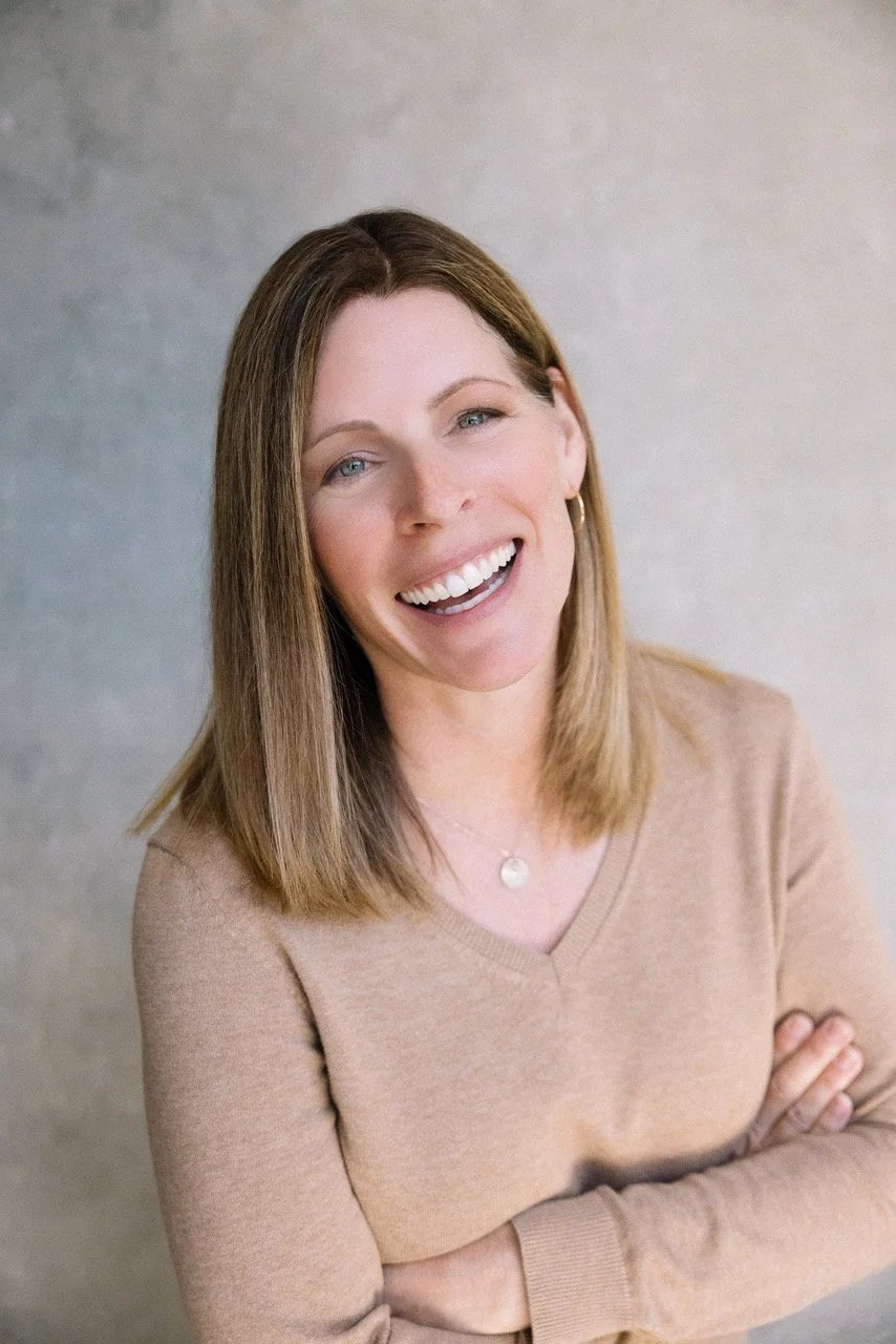 Dawn Mercado with shoulder-length light brown hair, wearing a beige sweater, gold hoop earrings, and a small necklace, standing with her arms crossed against a neutral background.