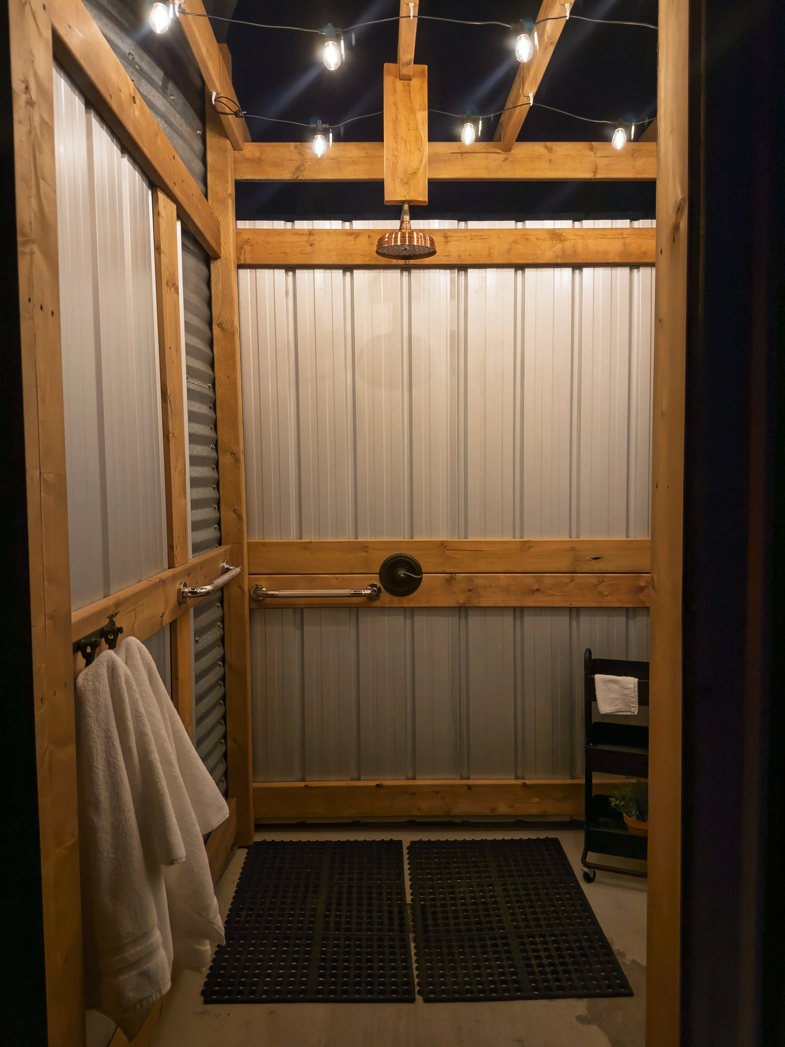Rustic outdoor shower with wooden frame, corrugated metal walls, overhead light bulbs, and a rain-style showerhead. Towels hang on a wall with hooks. Floor covered with black rubber mats.