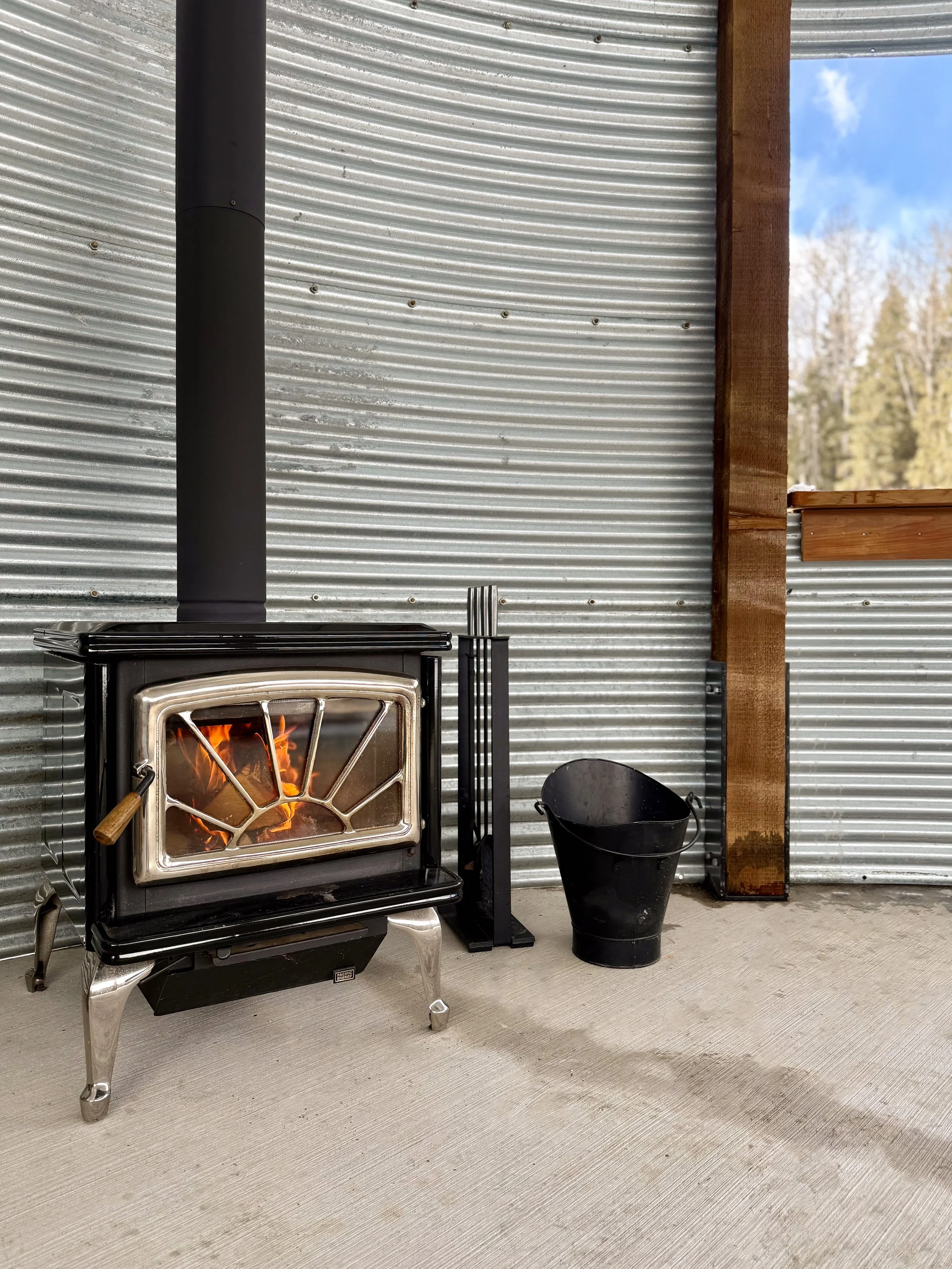 Wood-burning stove with chimney in reclaimed granary, next to a black metal bucket and indoor scene.