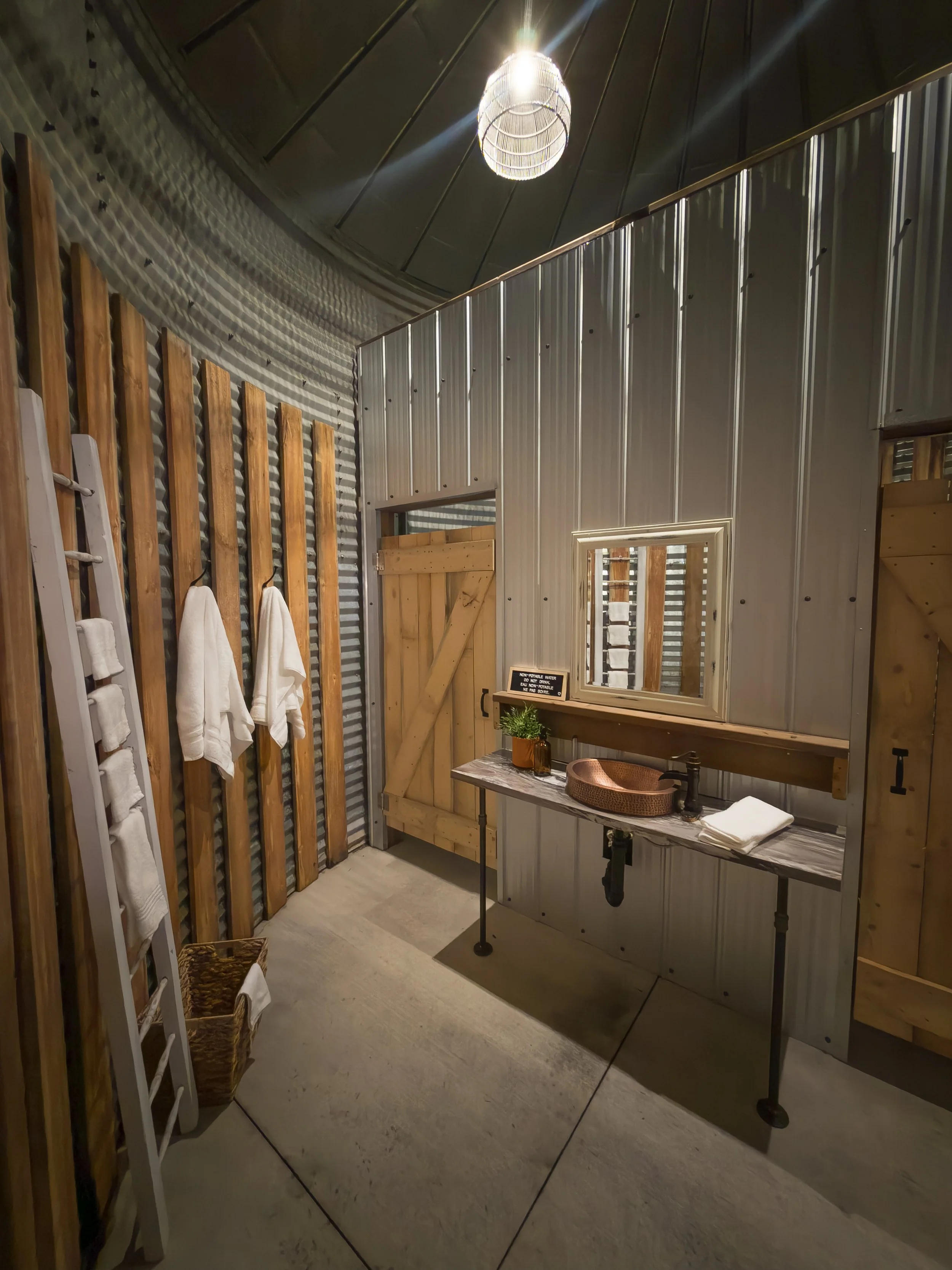 Modern rustic bathroom in reclaimed granary with corrugated metal and wood walls, copper sink, wooden countertop, towels on a ladder, wicker basket, and hanging light.