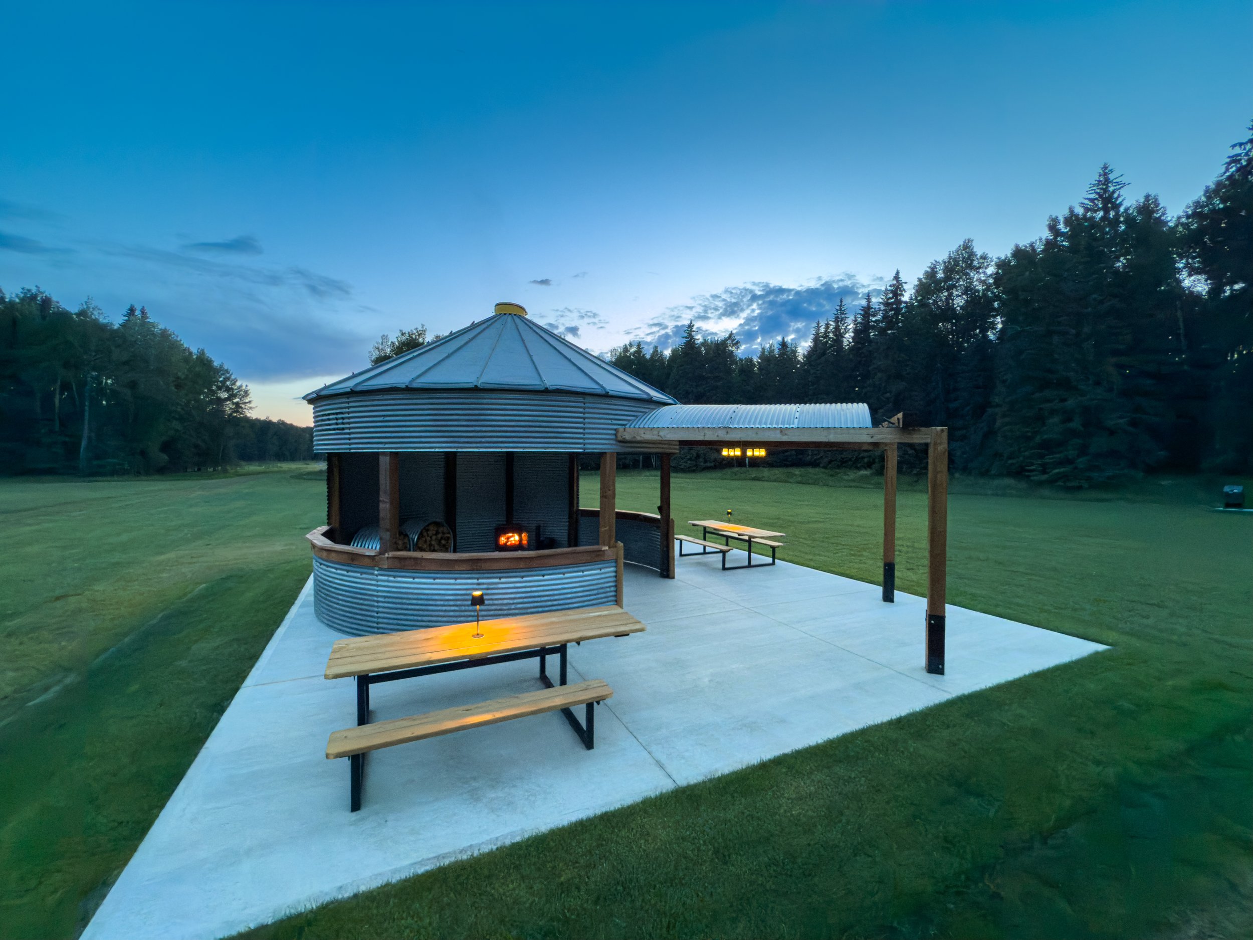 reclaimed granary sitting on a cement pad, mowed green grass, trees and lit fireplace at sunset